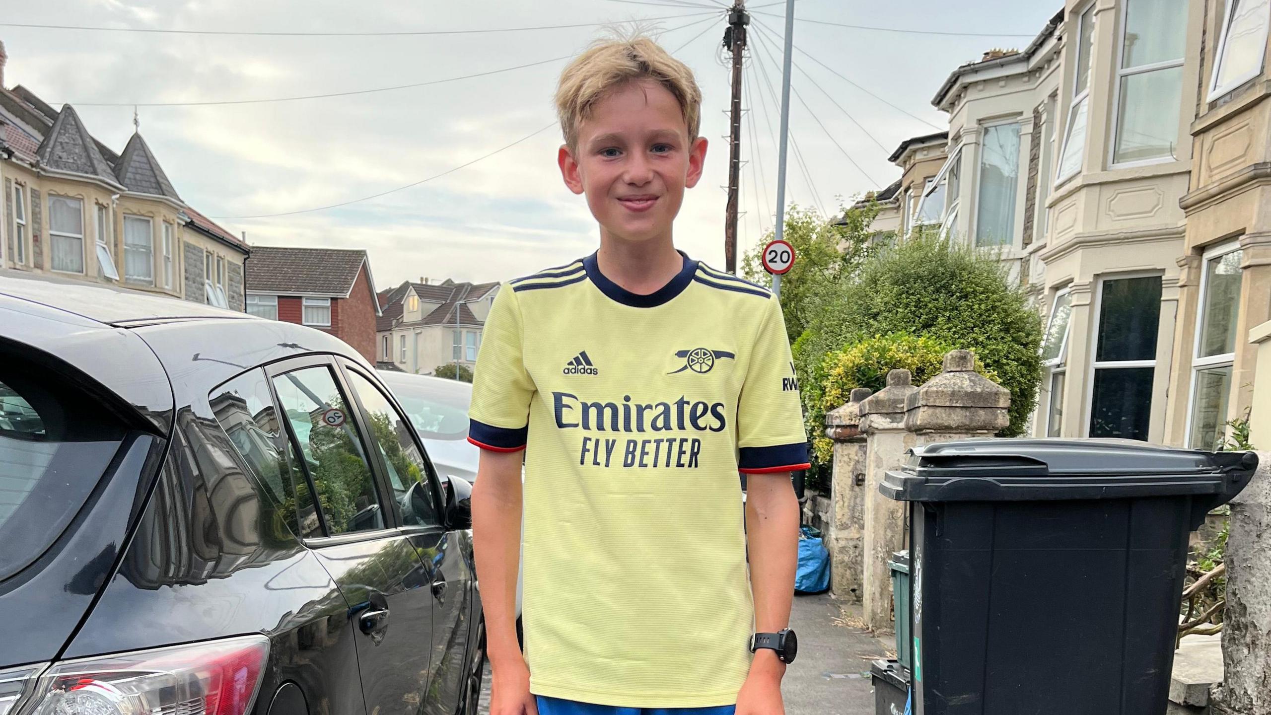 George stands in the street and smiles at the camera. He is wearing a light yellow Arsenal shirt.