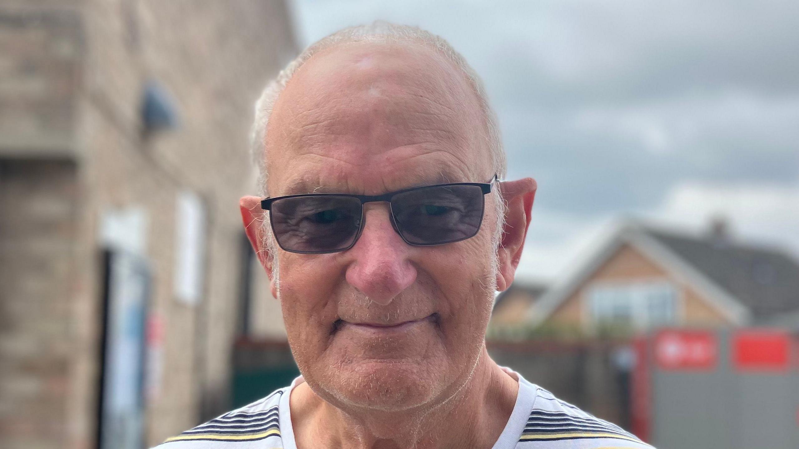 Roy Plowright smiles at the camera. He is largely bald with some thin hair on the side of his head. He wears sunglasses and a striped T-shirt. 