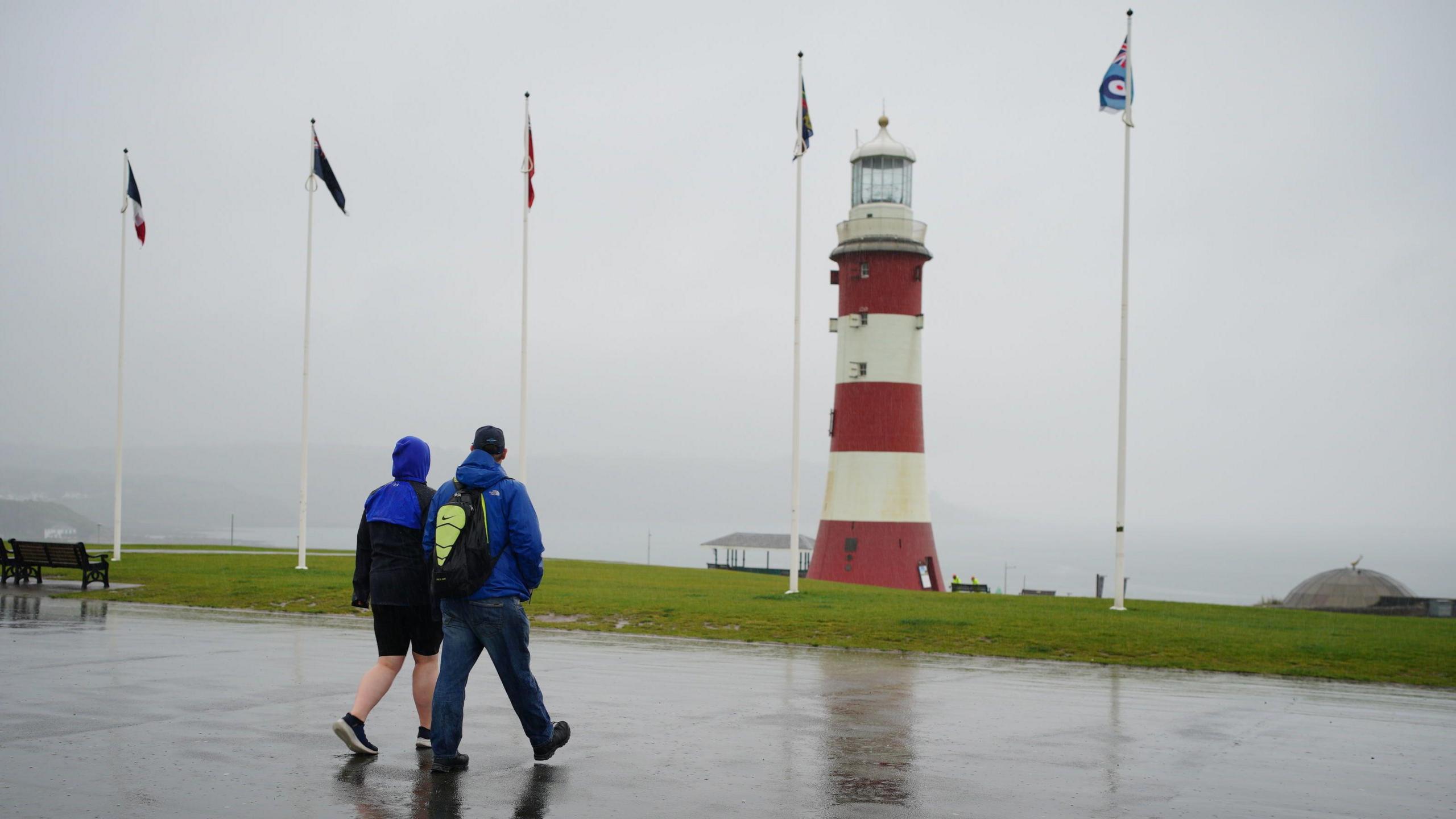 Two people in raincoats walking away from the camera with a lighthouse and flags in the background