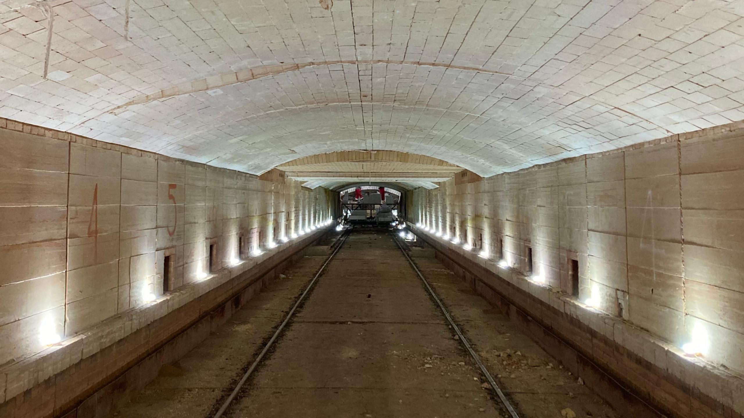 Interior view of a long industrial tunnel with a high, arched brick ceiling and parallel rails running down the centre. Small lights line both sides of the tunnel, illuminating pale stone walls marked with large painted numbers. At the far end, a piece of industrial equipment is visible on the tracks. The image is symmetrical, drawing the eye toward the distant vanishing point and highlighting the scale and length of the structure.