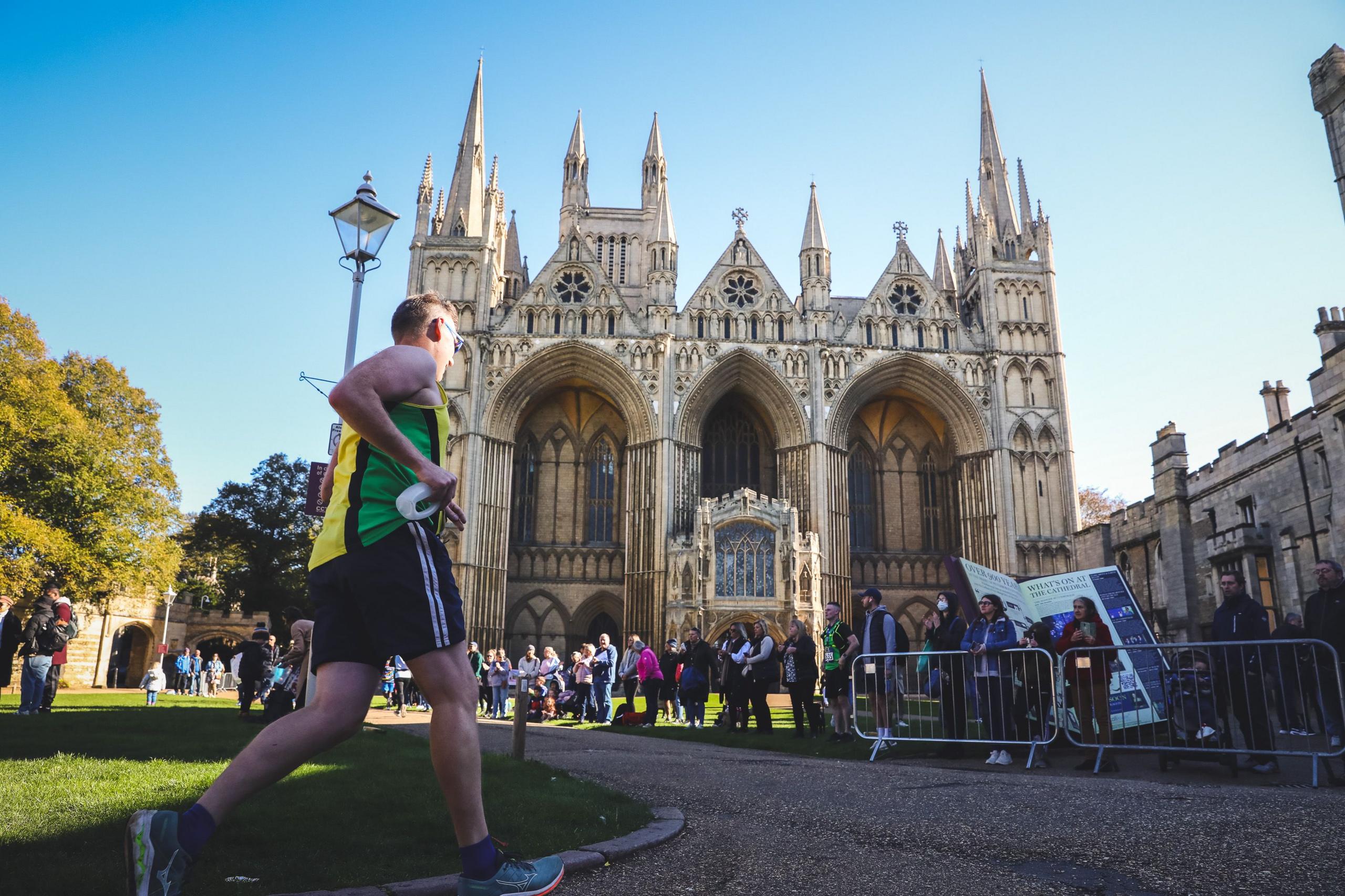 A runner in a yellow and green T-shirt  running towards Peterborough Cathedral- a tall medieval building with people waiting around it.