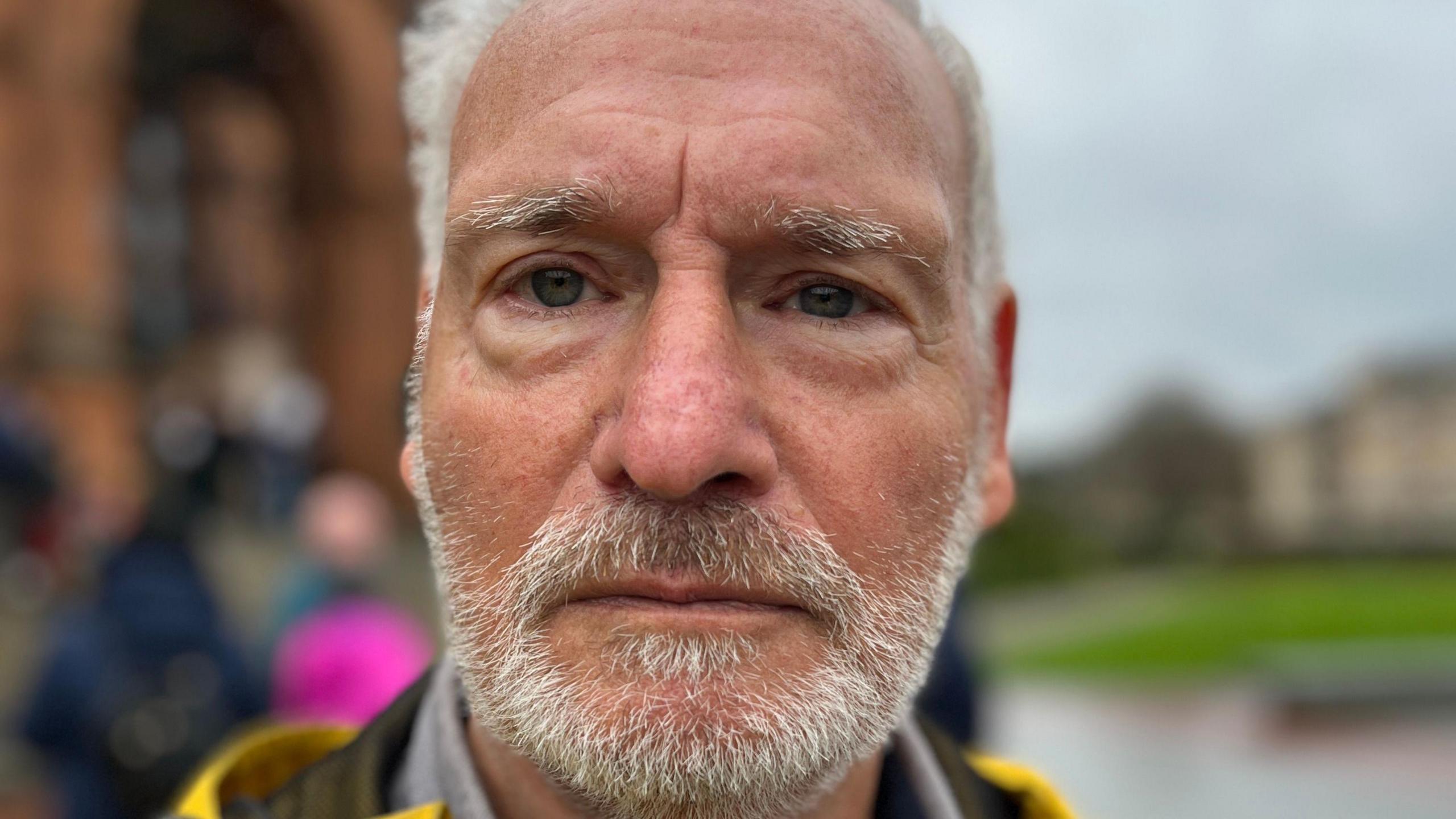 Sammy Stein, a man with grey hair and a grey beard. He is looking straight at the camera with a stern expression. The background is blurred.