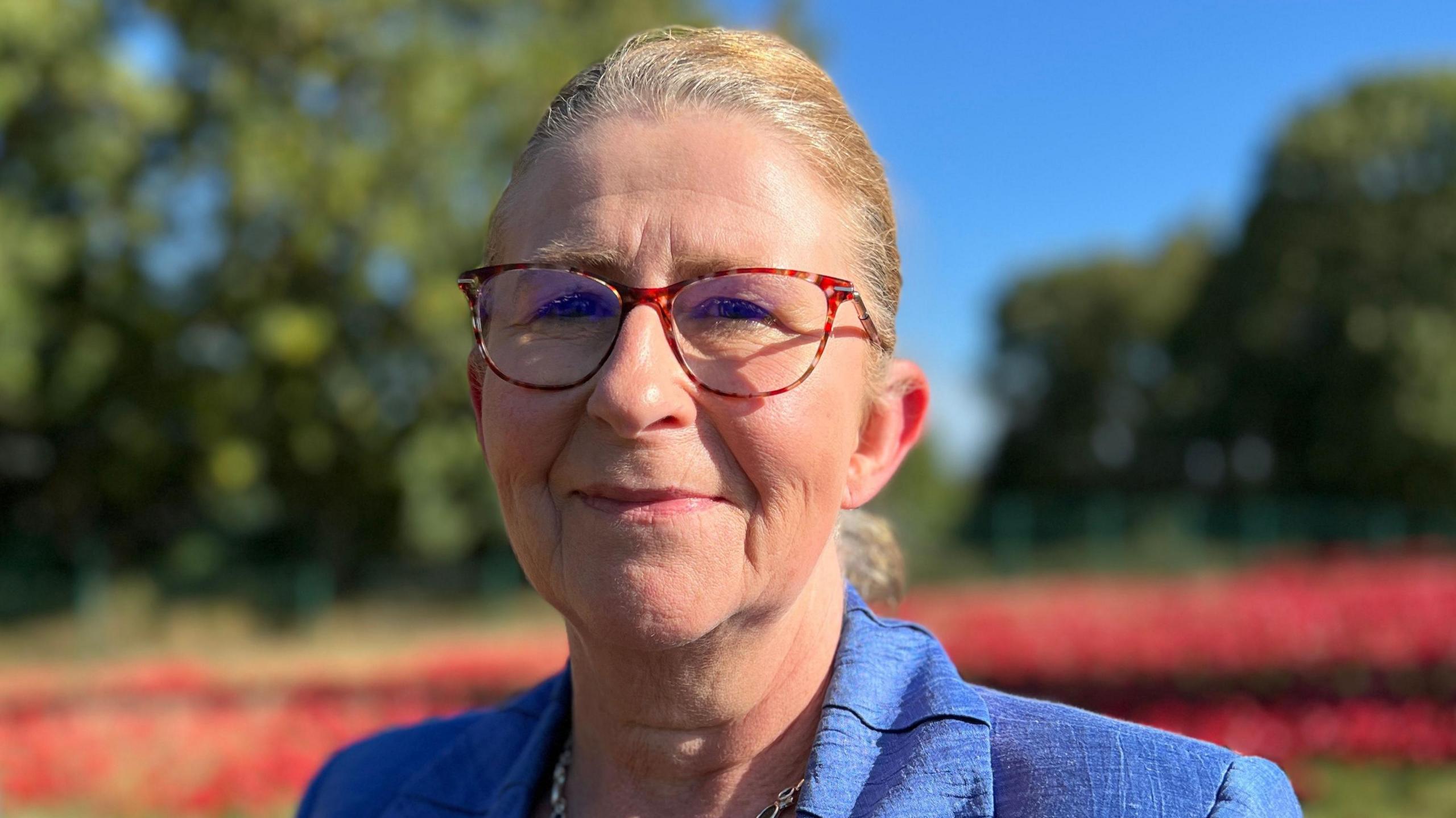 Nicky van der Drift is standing in front of a sea of ceramic red handcrafted poppies. Nicky is wearing red glasses, has blond hai and a blue blazer. The sky behind her is blue.