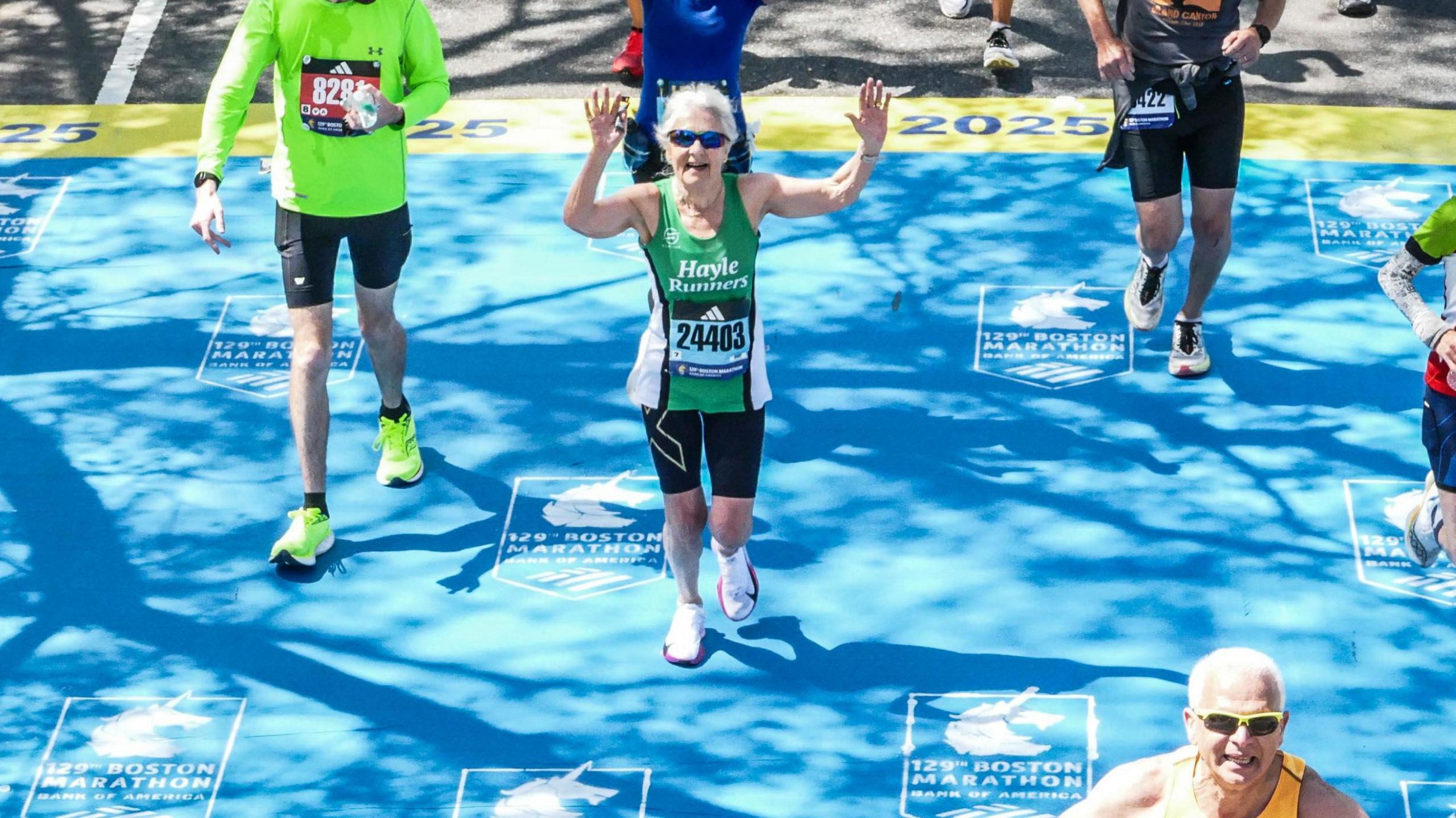 Andrea Simmons waves her arms as she crosses the finish line at the Boston Marathon. A blue mat with the Boston Marathon logo is on the floor. Several other runners are also crossing the finish line.