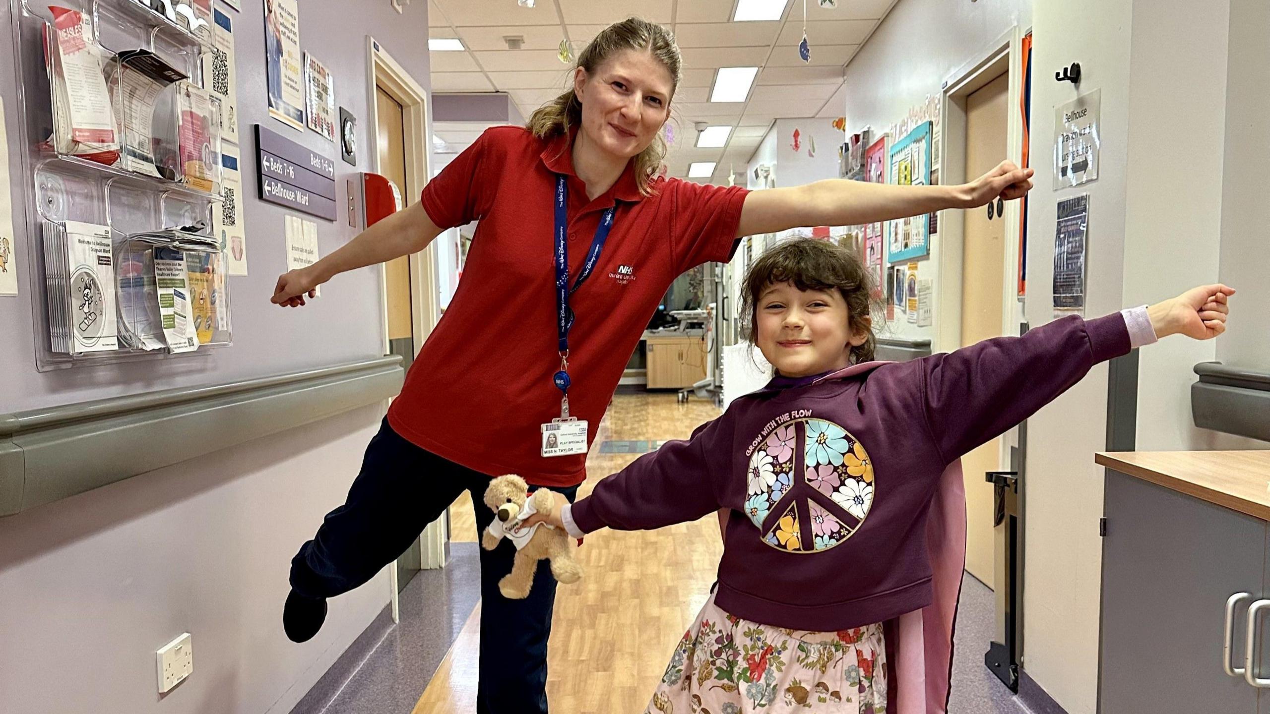 Sylvie with Play Specialist Naomi Taylor, smiling for the camera at the hospital while stretching their arms in a superhero flying pose. Sylvie is holding a small beige teddy bear.