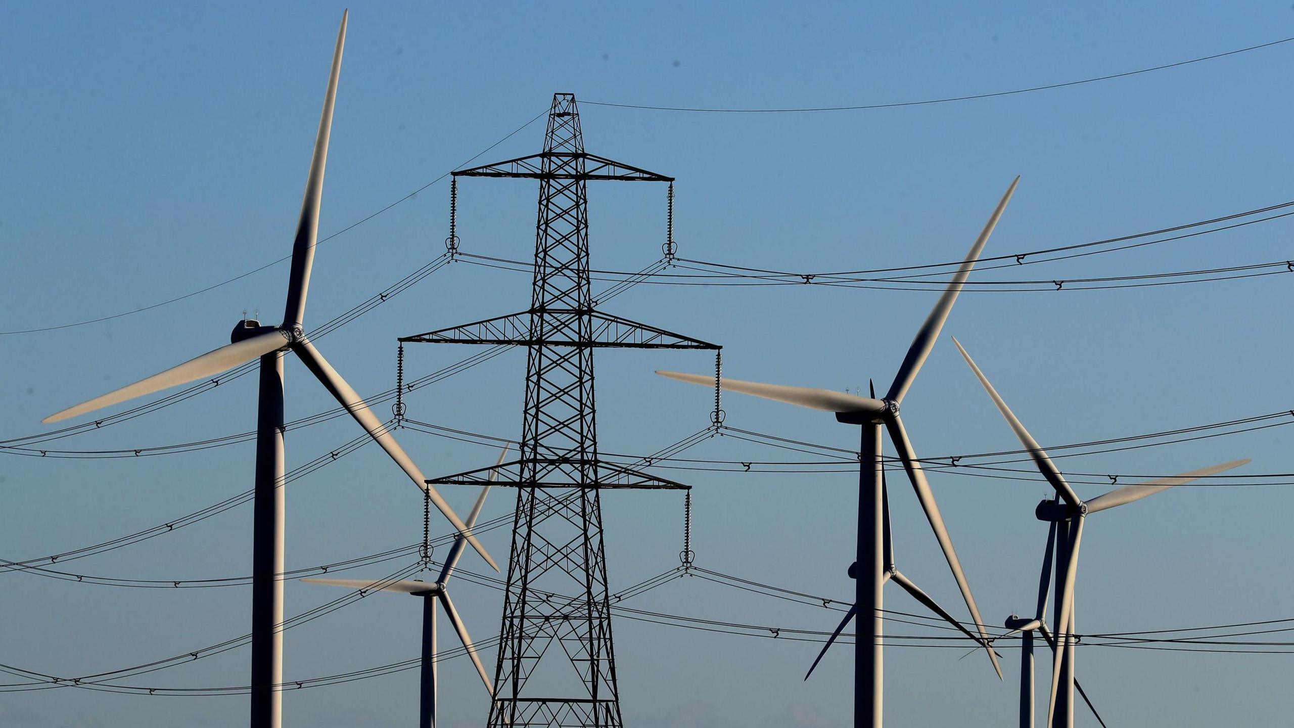 A group of five wind turbine, in shadow against a blue sky, stand in front of a large electricity pylon with cables stretching to either side.