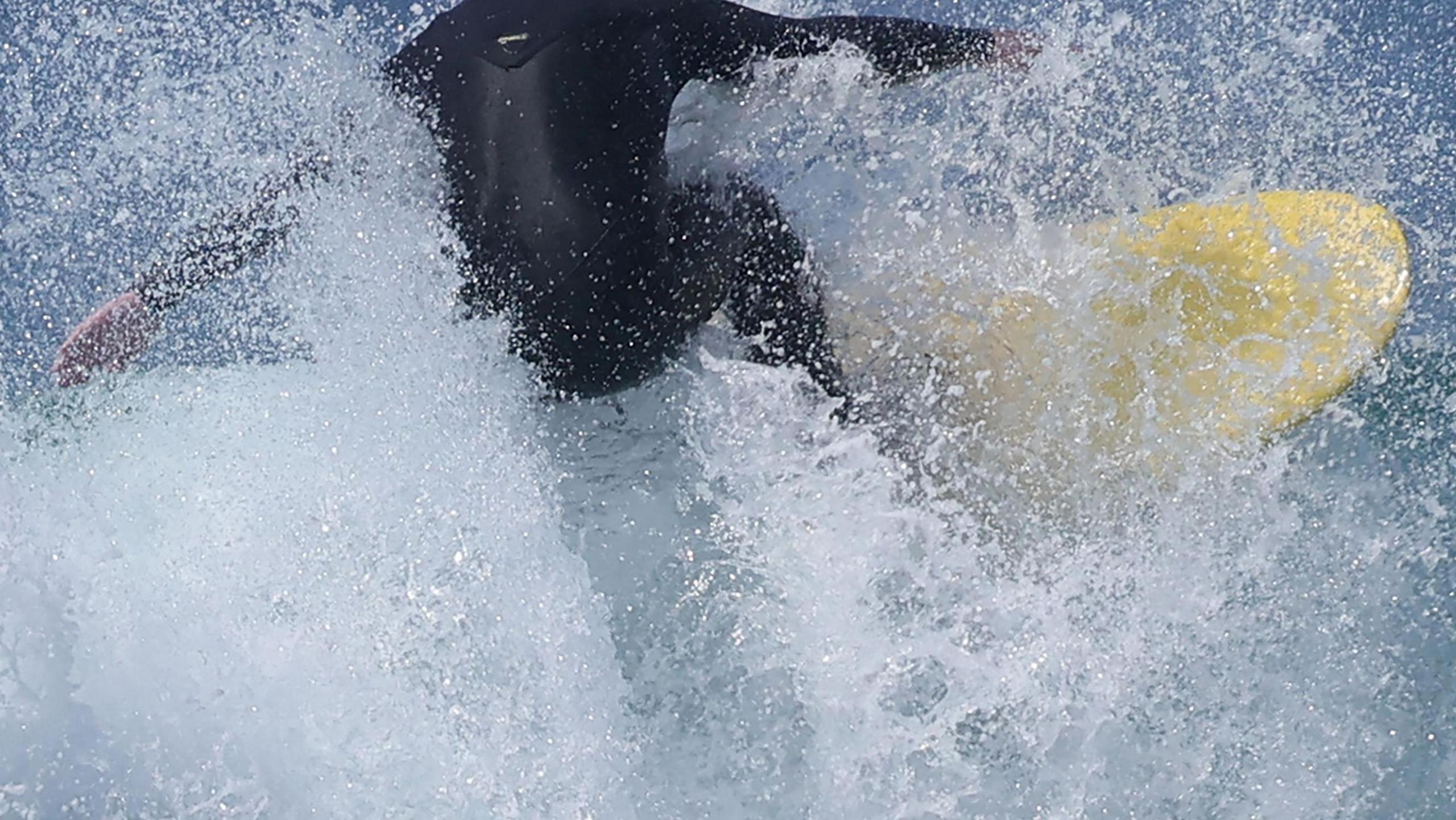 A surfer crashes through the waves on a yellow surfboard. He is wearing a black wetsuit.