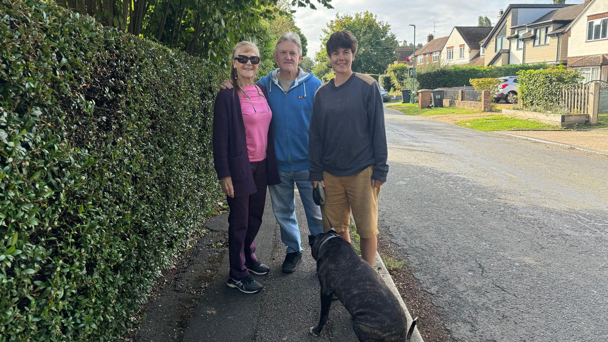 From left to right: Hilary Sewter, Clive Sewter and Alice Cherry. There is also a black dog in the picture. They are standing next to a hedge on Potters Lane. The road and some houses are in the background.