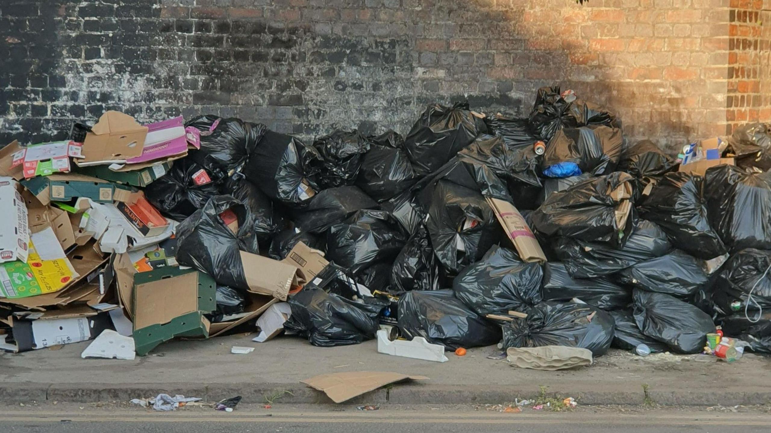 Piles of black bin bags, cardboard and other litter against a dark wall near a kerbside. There are about 30 bin bags in the picture.