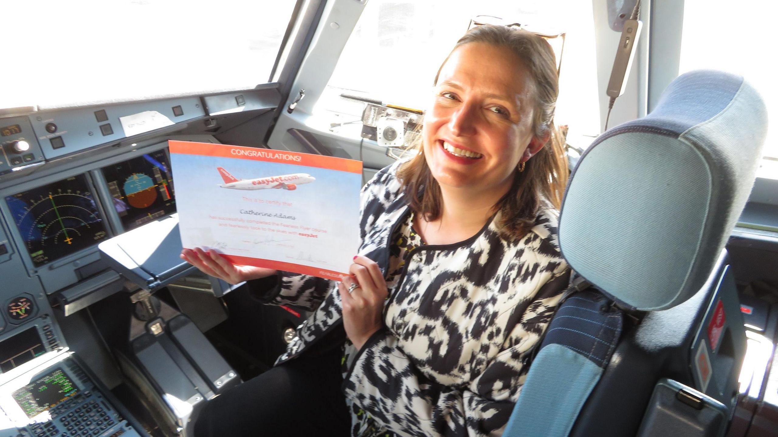 Cathy sits in the cockpit of an Easyjet plane holding a certificate that is orange and blue. It says CONGRATULATIONS at the top. She is smiling at the camera and wears glasses on her head, and a black and white jacket and black trousers.