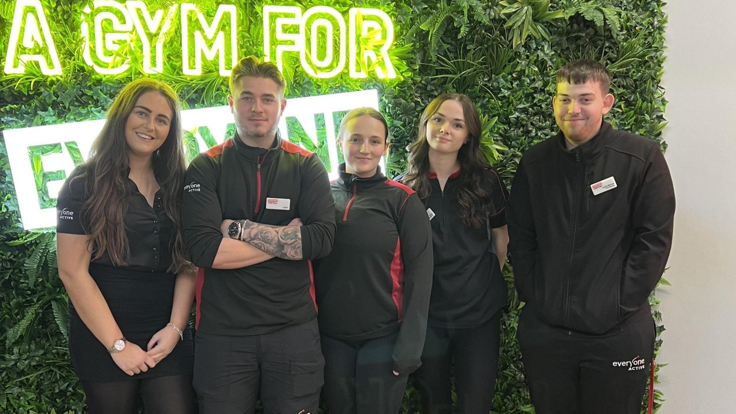 A group of young gym staff, all wearing black uniforms, pose for the camera inside Horfield Leisure Centre. Behind them is a wall of fake greenery with a neon sign in it