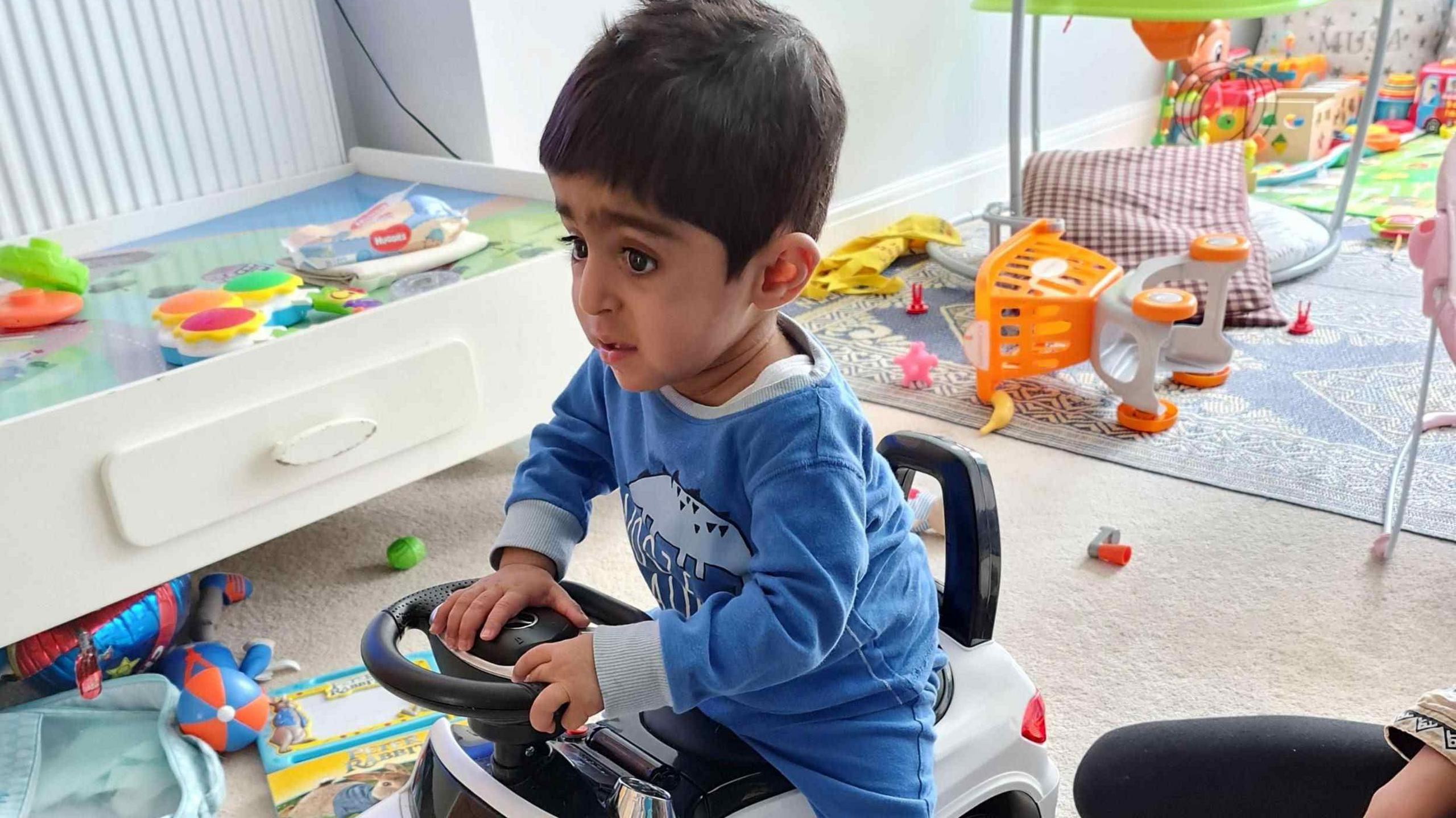 Eisa at home in a playroom, surrounded by toys is riding a play car. He is wearing a blue baby grow, has dark hair and eyes.