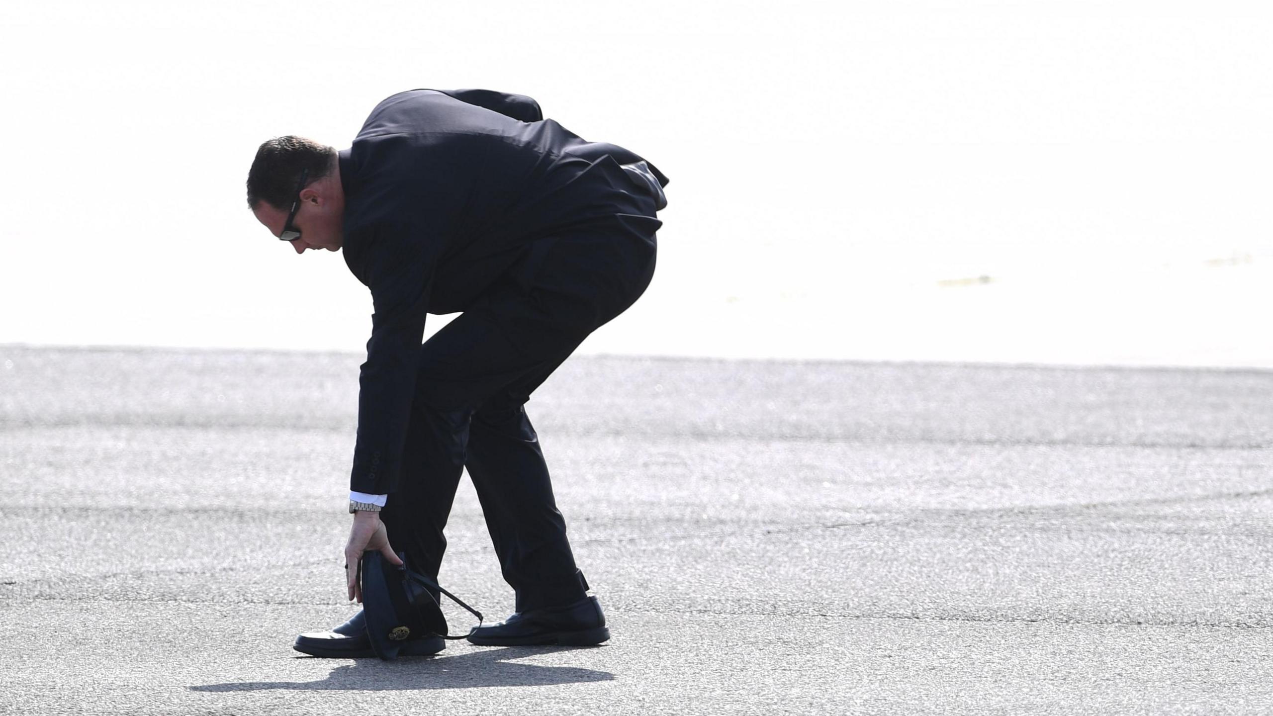 A suited security officer wearing sunglasses bends over to pick up a dark hat from the tarmac.