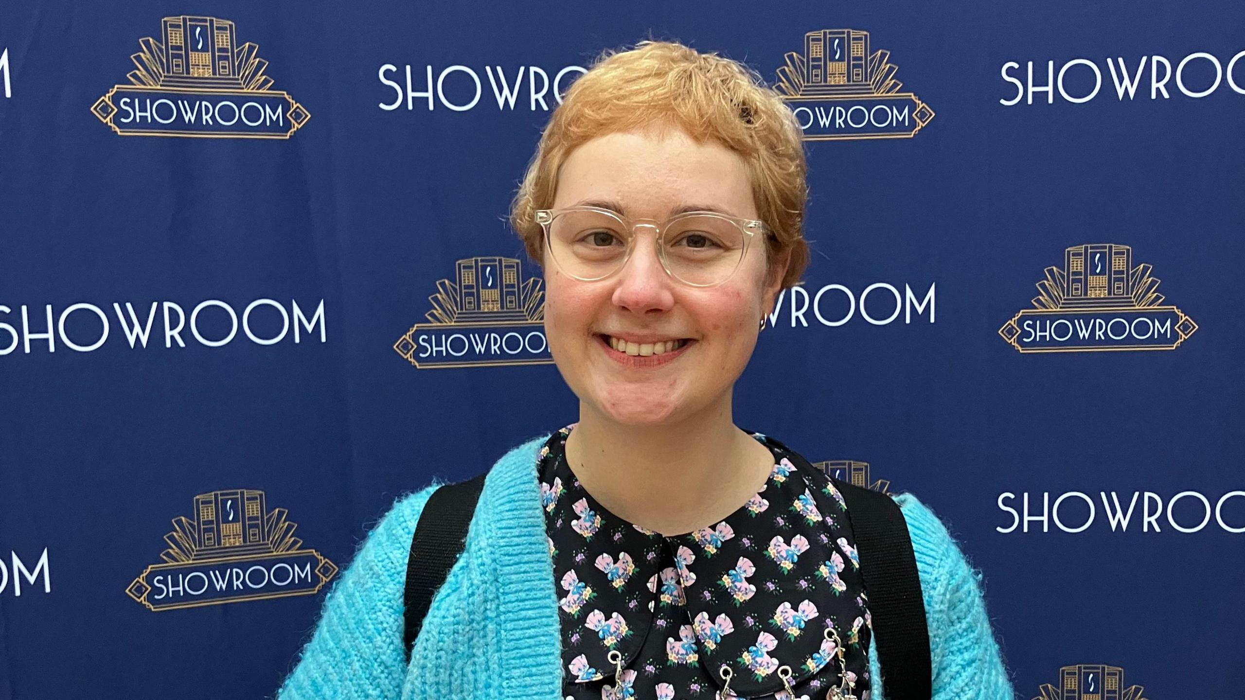 A woman with glasses and short blonde hair. She is wearing a light blue cardigan and black dress with pink cats on. Behind her a dark blue board reads: Showroom. 