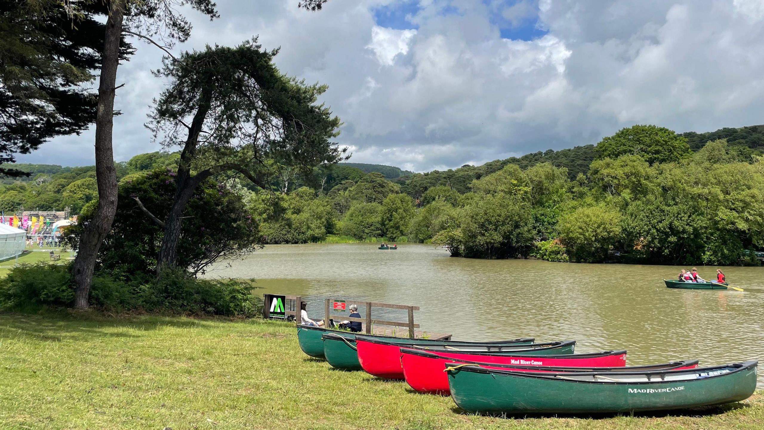 Canoes in red and green line a lake bank. A paddleboat can be seen out on the lake with a group of people in. Trees line the opposite bank of the lake.