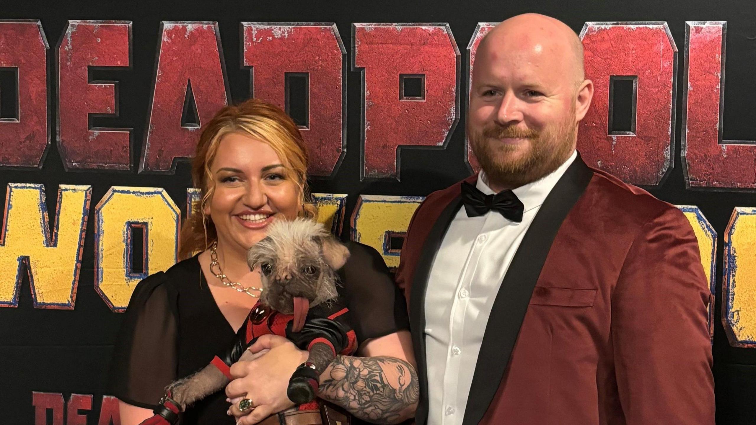 A smiling Holly Middleton, who wears a black dress, gold necklace and has tattoos on her arm, holds her dog Peggy, who is wearing a black and red costume, at an event to promote Deadpool and Wolverine. Her husband, wearing a burgundy dinner jacket, white shirt and black tie, stands next to her.