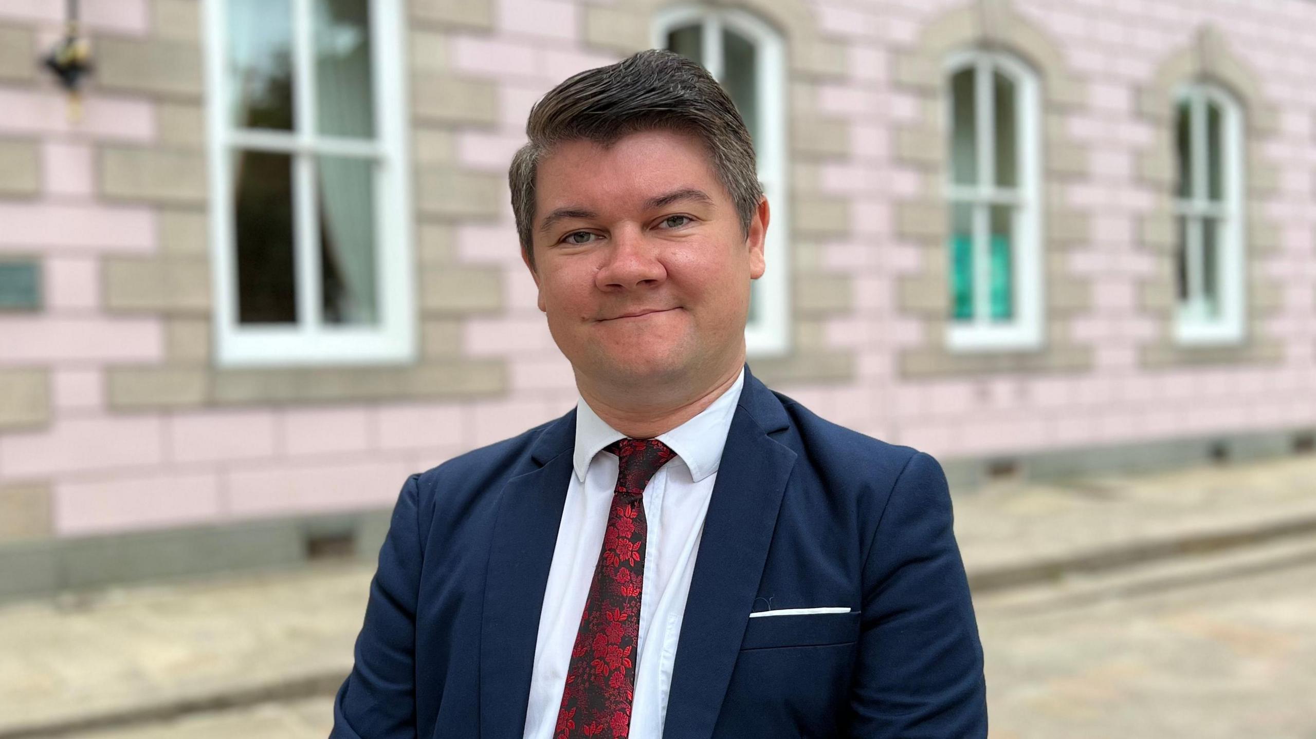 Deputy Sam Mézec standing in the Royal Square in front of the States Assembly building. He is wearing a navy suit with a red tie.