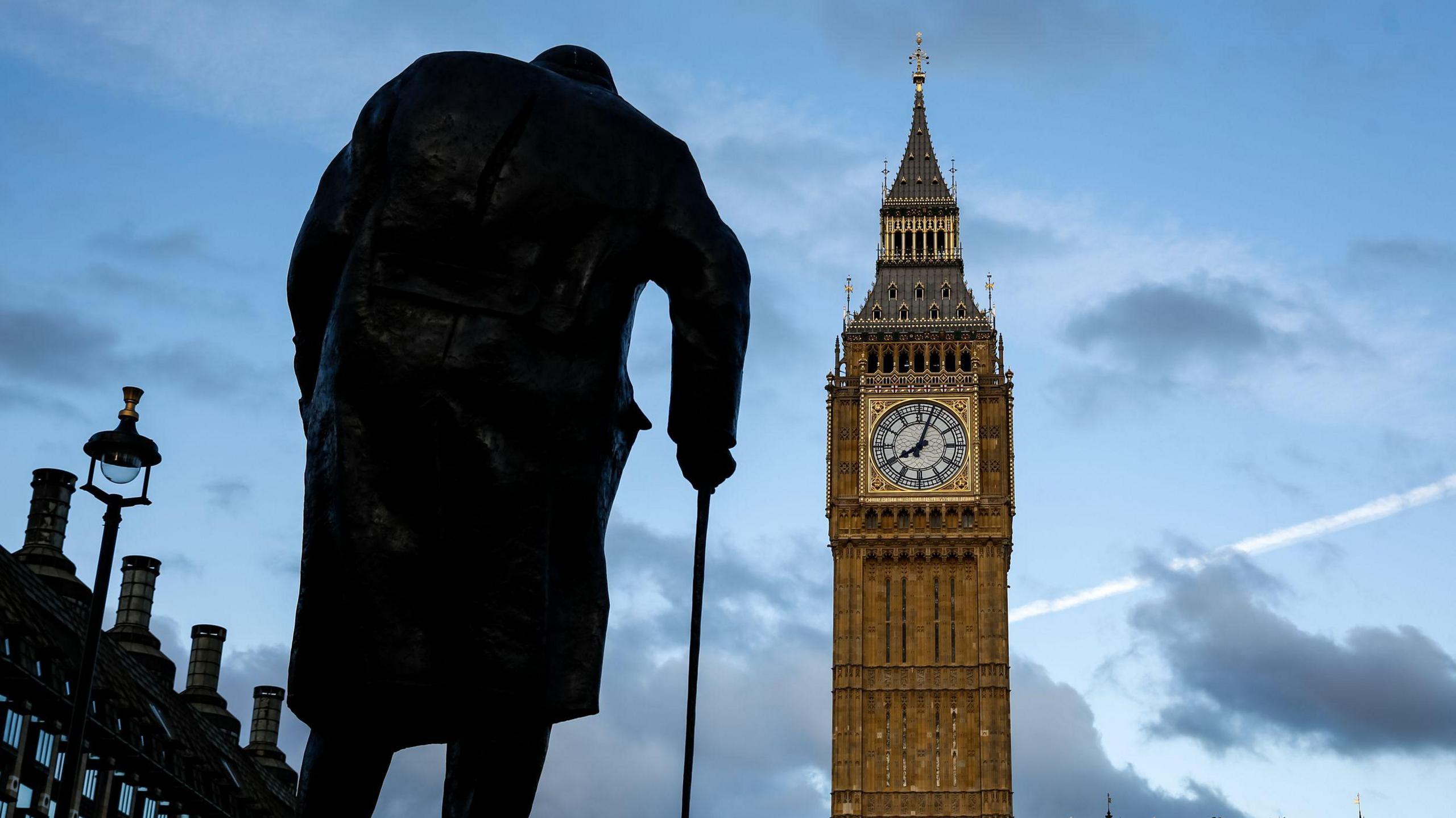 A Sir Winston Churchill statue seen in front of Big Ben in central London. The stature is in silhouette along with some buildings to the left, and Big Ben is in full colour. The picture was likely taken at dusk as the sky is beginning to go dark.