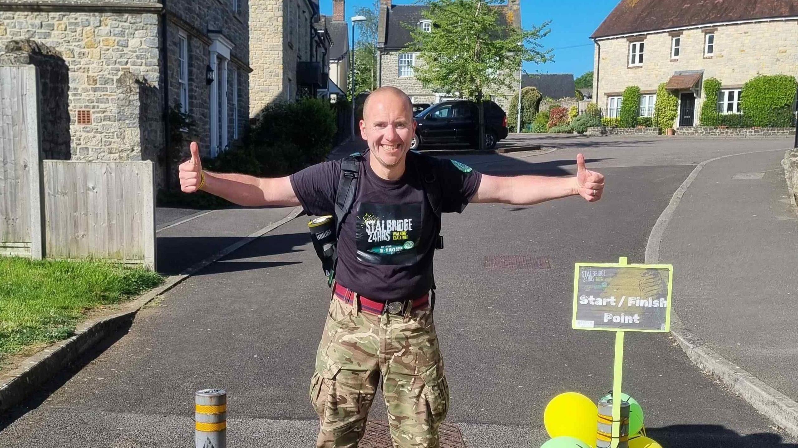 Martyn Harris is standing with thumbs up and smiling at the camera, he is wearing a black t-shirt advertising his Stalbridge 24 hours walking challenge and army trousers.
