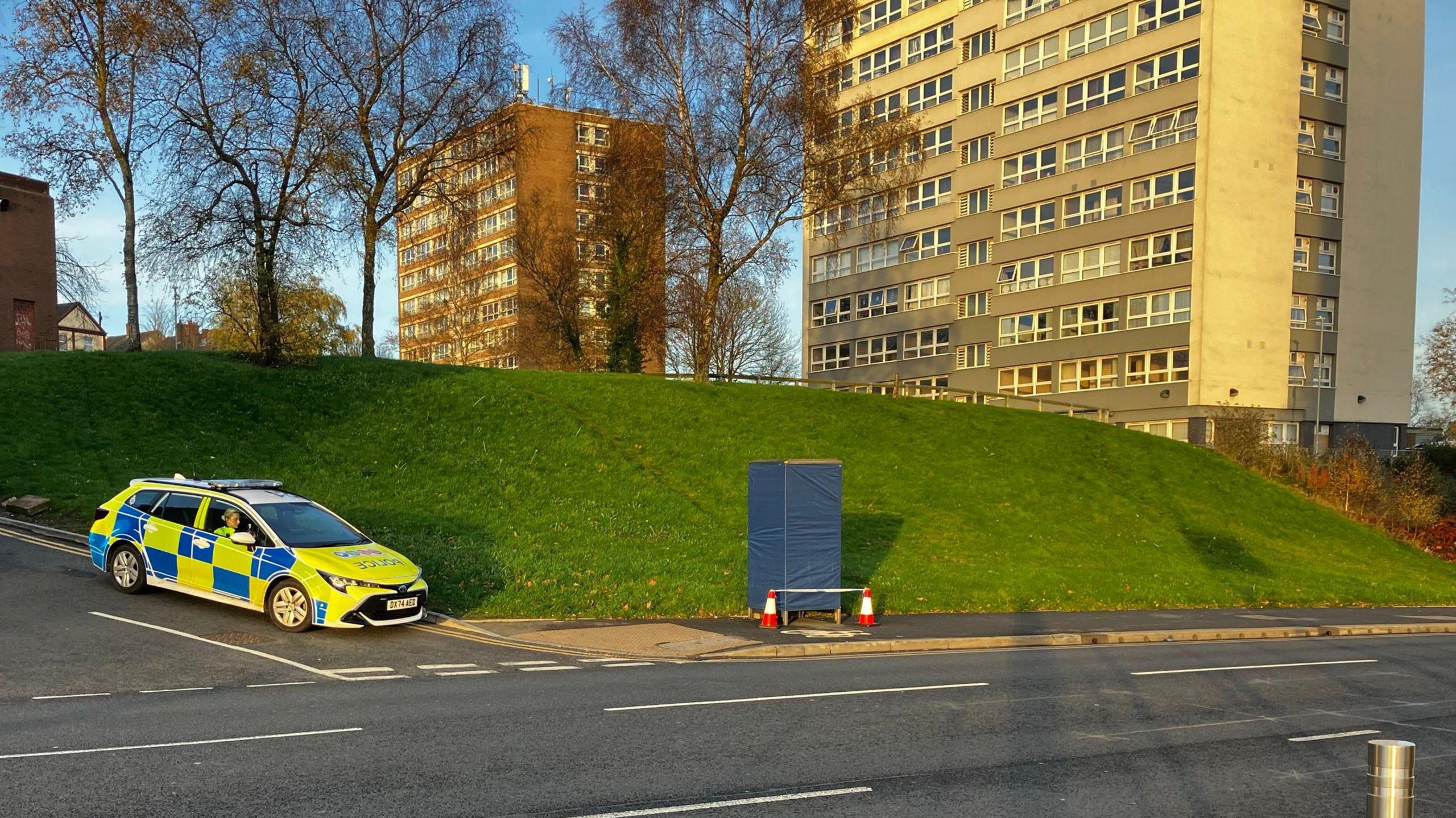 A police car parked at the side of the road at a junction. A phone box on the pavement next to the road is covered in blue tarpaulin with two cones in front of it, which are connected with blue and white police tape. Behind the phone box is a small green hill with trees at the top and tower blocks in the background.