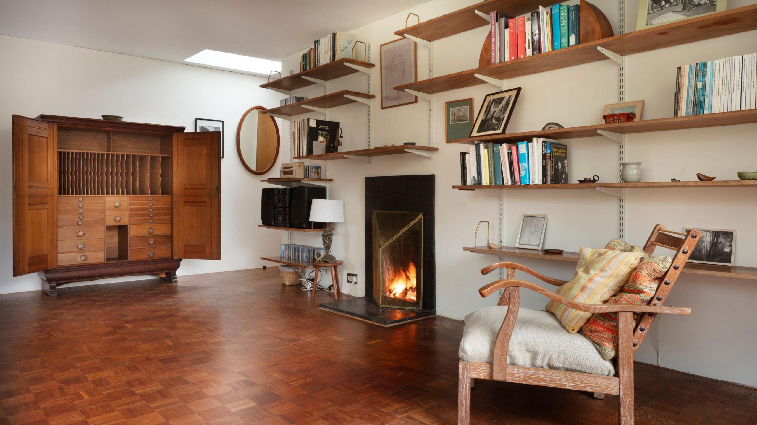 A view inside Holst's living area inside her home. A fireplace can be seen as well as a nearby wooden chair. A large oak cupboard also sits upon one of the room's walls with its doors open. Bookshelves line the walls.