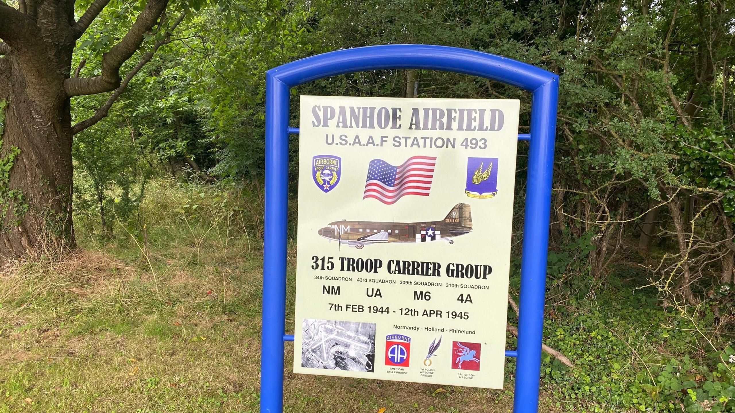 A sign about Spanhoe Airfield explaining it was a World War Two USAAF station held in a bright blue frame with trees behind