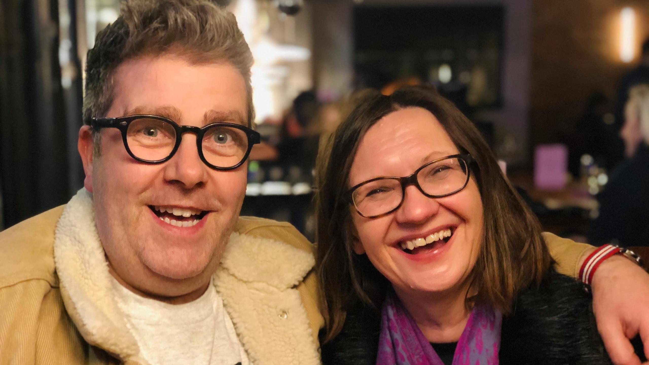 David Burgess has his arm around Sandra O'Hagan's shoulder. Both are smiling. He is wearing a cream cord jacket with fur-lined collar and a grey T-shirt and she is wearing a dark top with a cerise scarf.