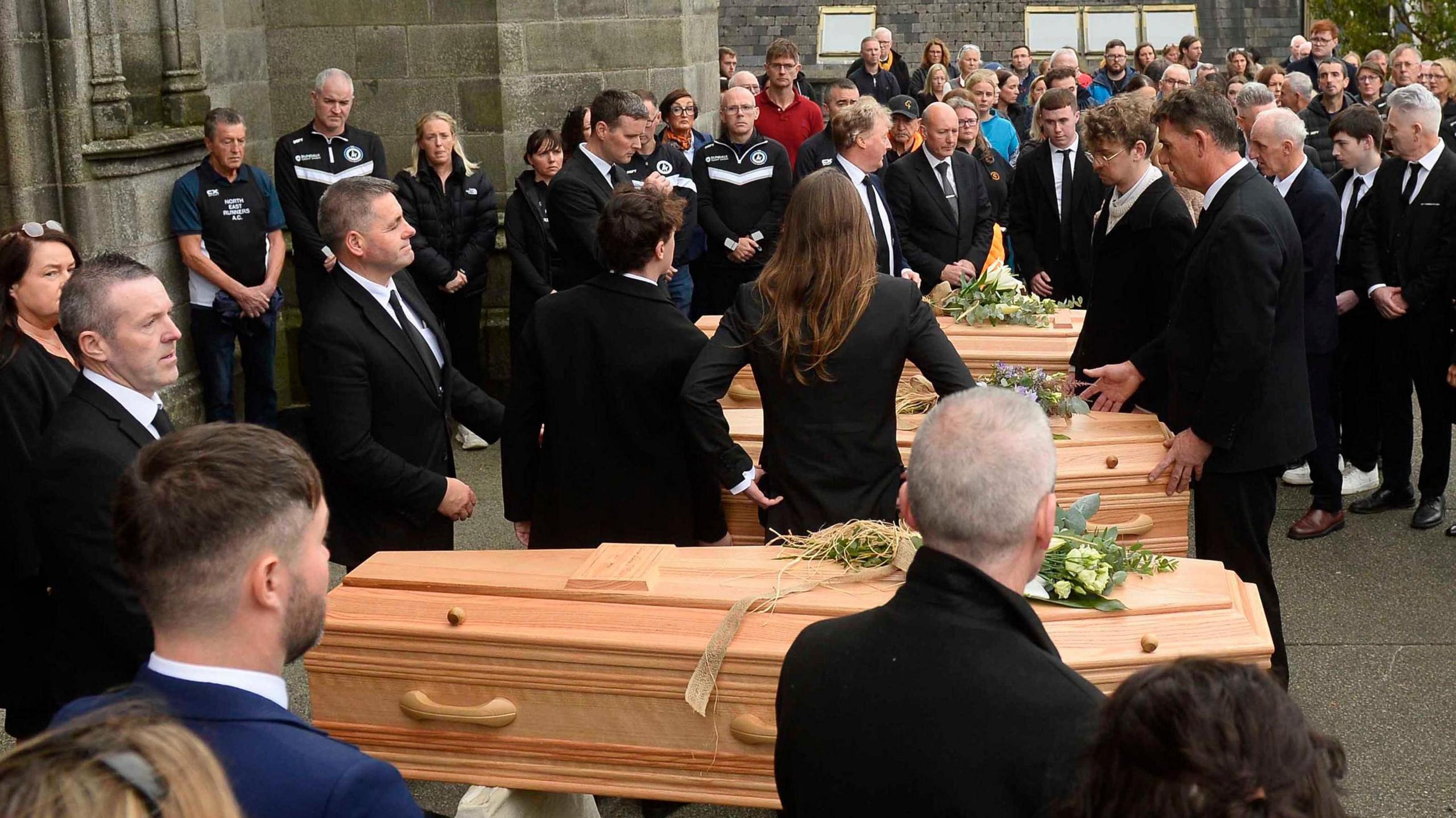 Three coffins in a row outside a church. There are flowers on each of them and people all around them, dressed in black.