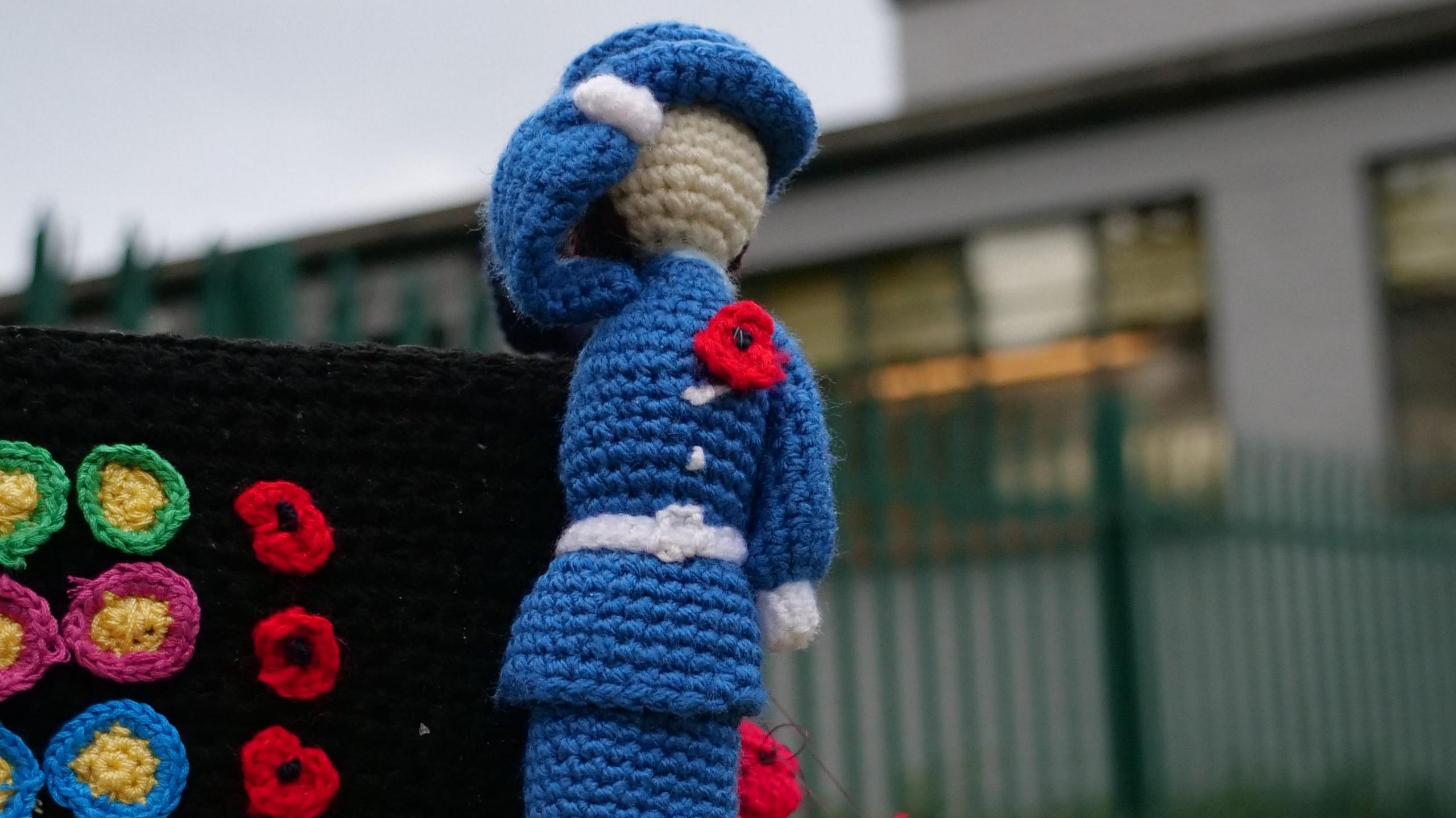 A knitted figure of a woman in naval uniform, wearing a poppy and saluting, next to a knitted recreation of a WW2 Bombe machine