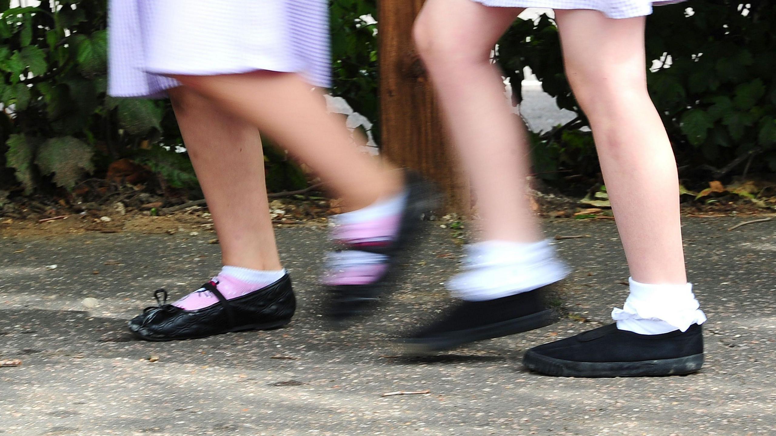 A generic view of the lower half of two anonymous primary school pupils as they walk. They wear school summer dresses with black shoes and socks.