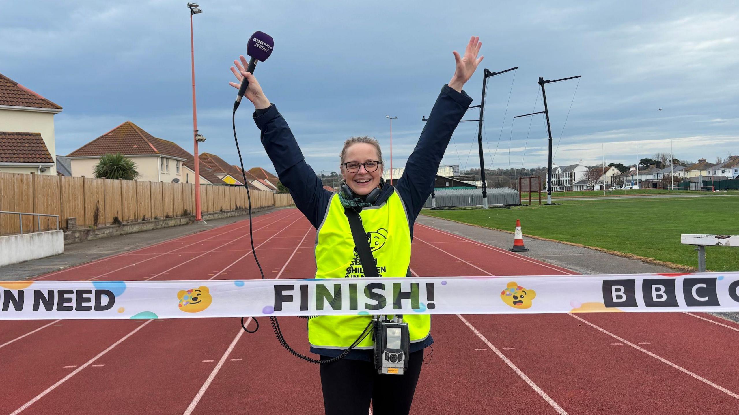 Jenny Mullin crossing the finish line. She is wearing a green vest and walking on a red multi-lane running track towards a line which says "finish" on it