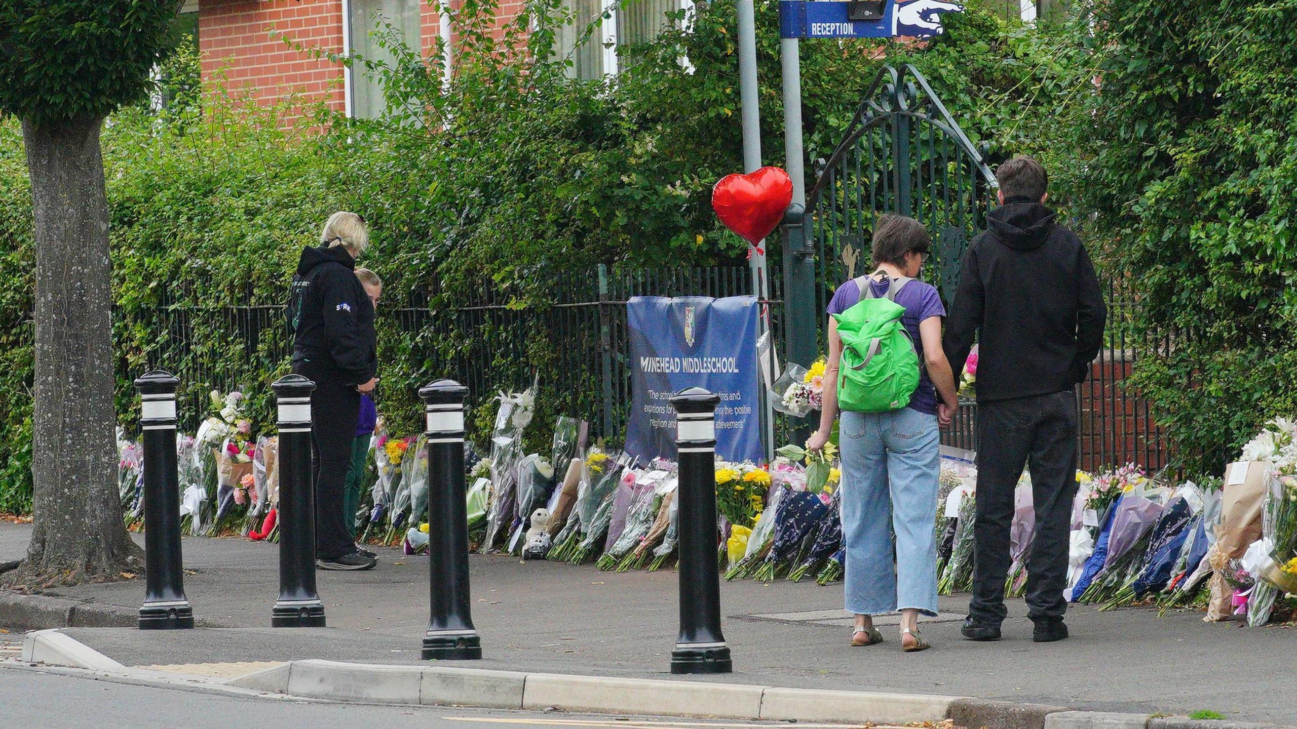 Four people stand with their backs turned outside Minehead Middle School. They are looking at bunch of flowers which have been lined up against the metal fence outside the school. There is a red balloon in the shape of a heart attached to the fence.
