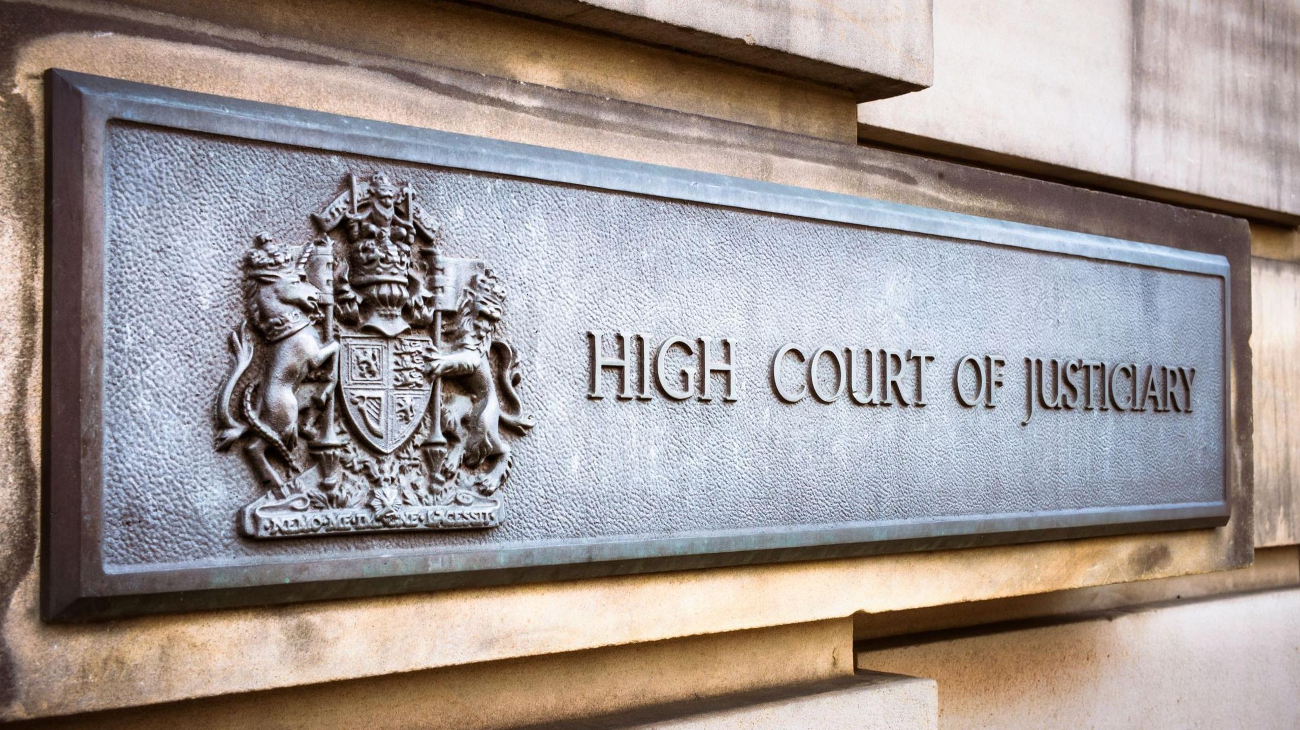 A grey metal sign on the sandstone block exterior wall of the High Court in Edinburgh. The sign has a royal crest and the words: "High Court of Justiciary".