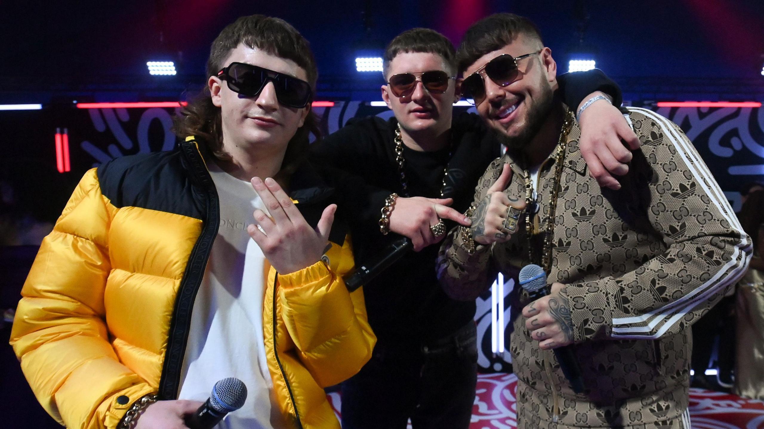 The three members of Bad Boy Chiller Crew are wearing dark sunglasses and facing the camera while holding microphones. One is in a bright yellow and black coat on top of a white t-shirt. Another is dressed in black with a gold chain around his neck and wrist. The other is in a patterned beige tracksuit. They are gesturing with their hands and are stood on a stage, lit by neon lights.