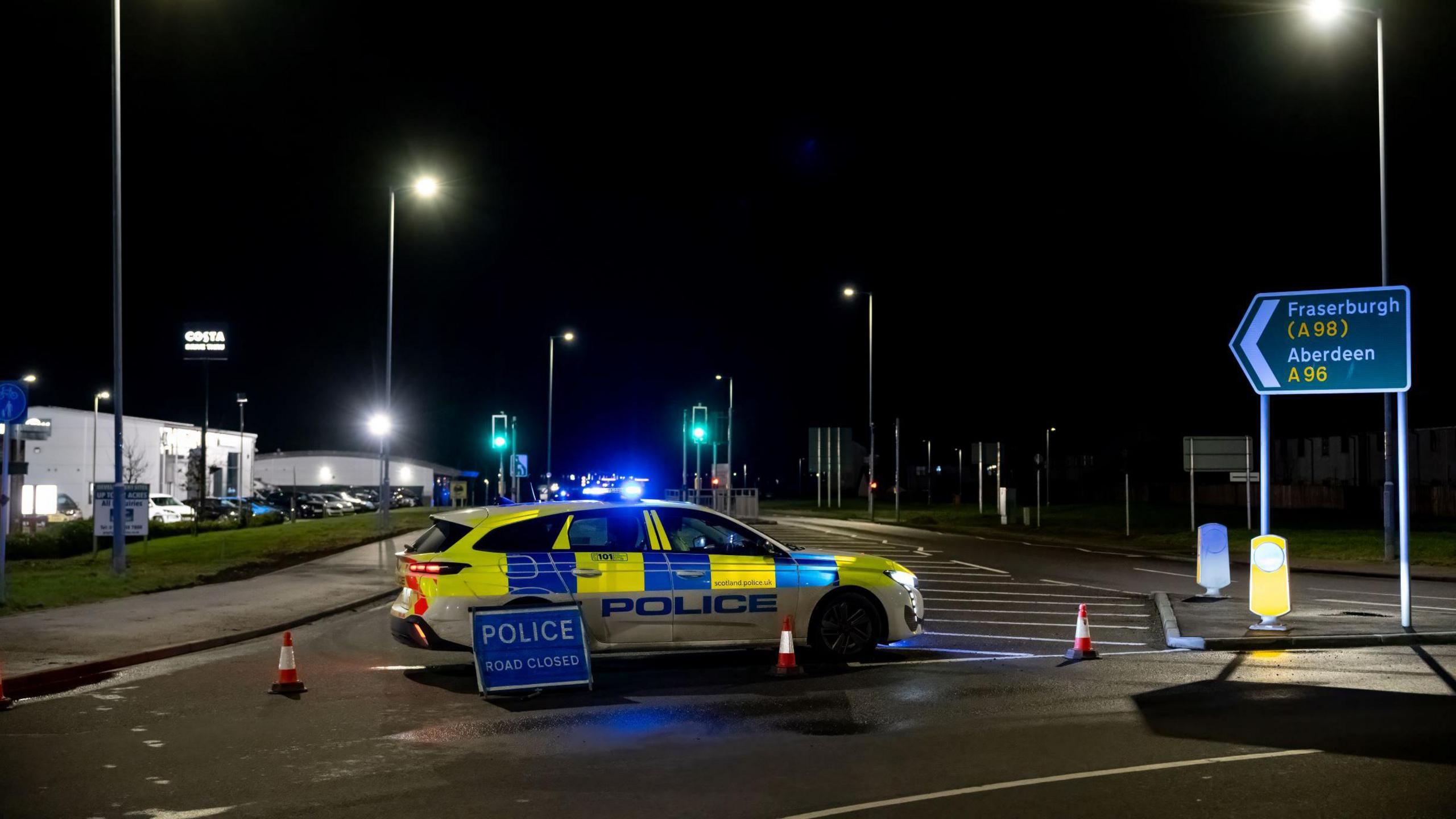 A police car parked across a road. There is a blue accident sign in front of the car. Several cones are in a line across the road. The police car has its blue lights activated.