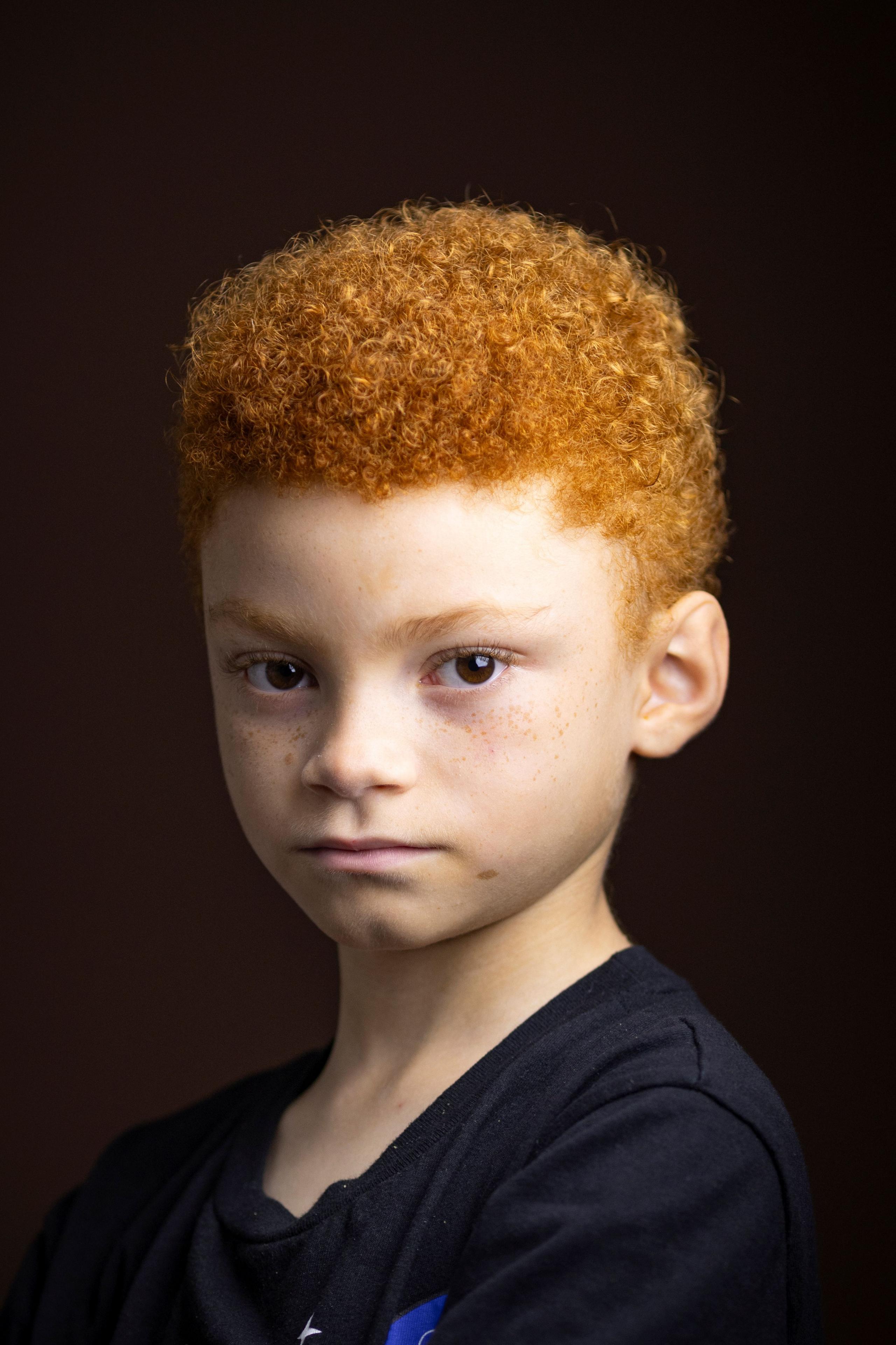Close-up studio portrait of a child with short, tightly curled red hair and freckles, wearing a dark T-shirt. The child faces the camera with the head turned slightly to one side against a dark brown background. Soft, warm lighting highlights the hair texture and facial features.