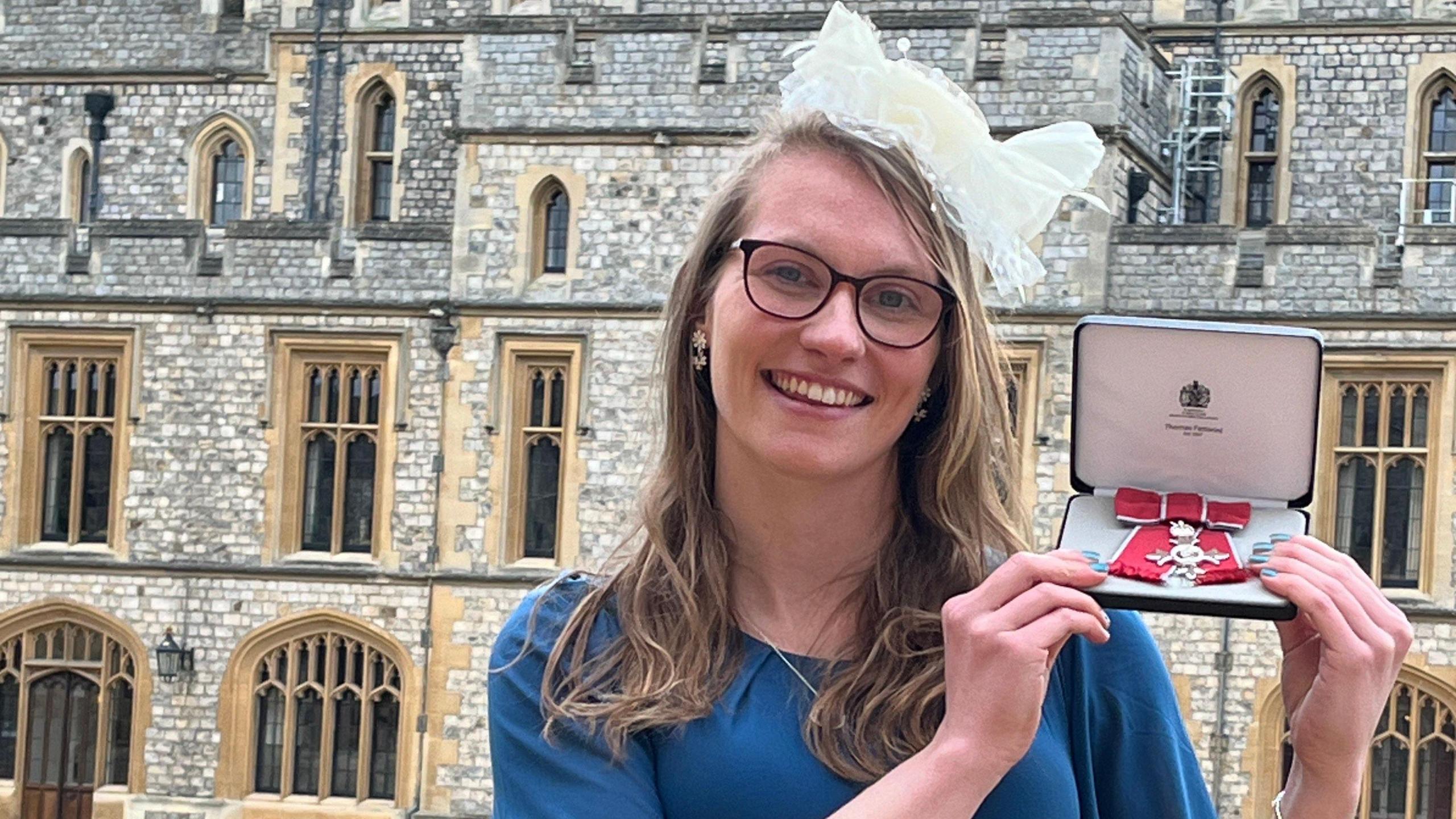 A woman  with shoulder length blonde hair is wearing round black framed glasses, a cream fascinator and a blue dress. She is smiling while holding a box with her MBE award medal inside.