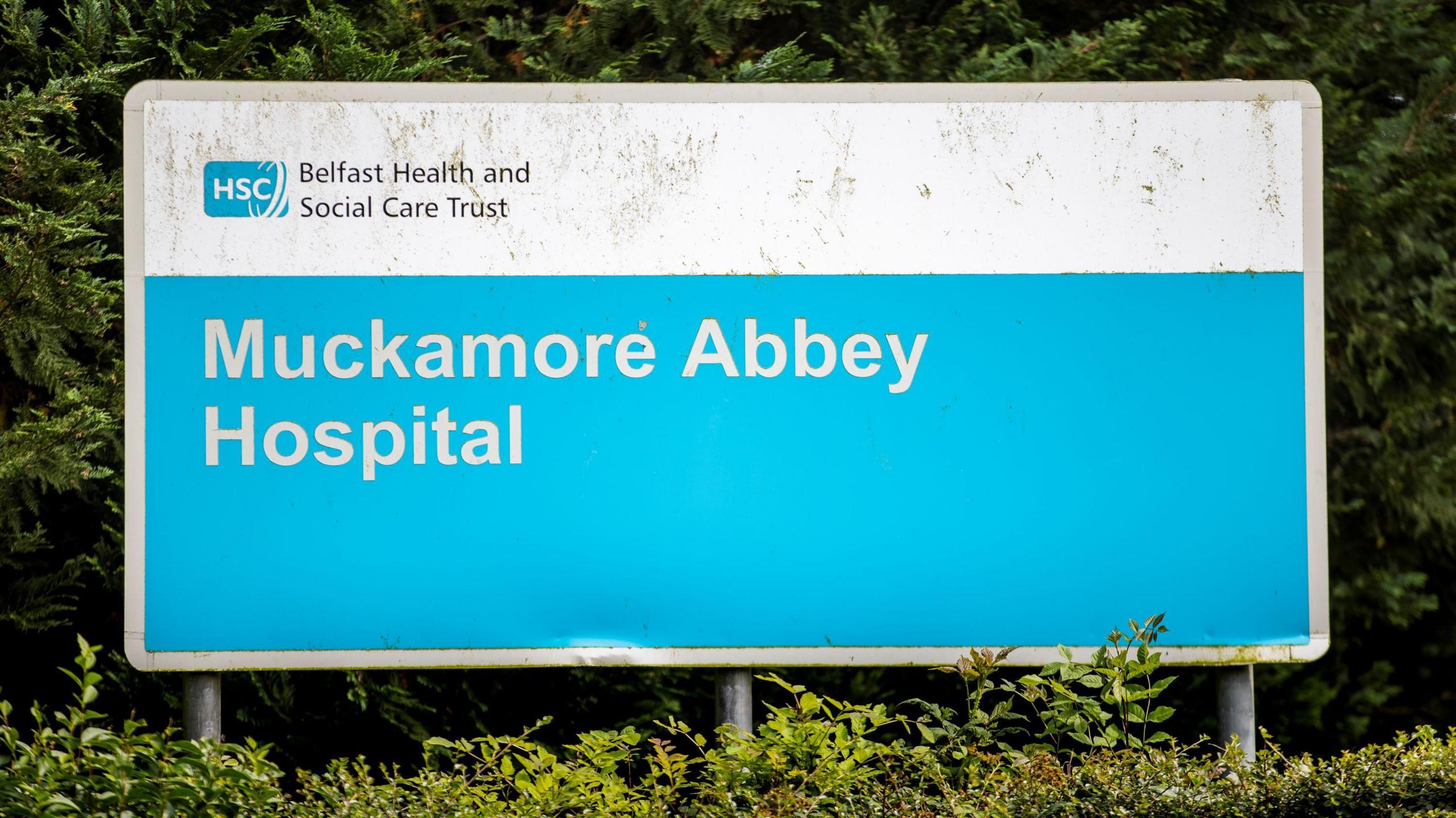 A blue and white sign that says Muckamore Abbey Hospital. It sits amongst a green bush.