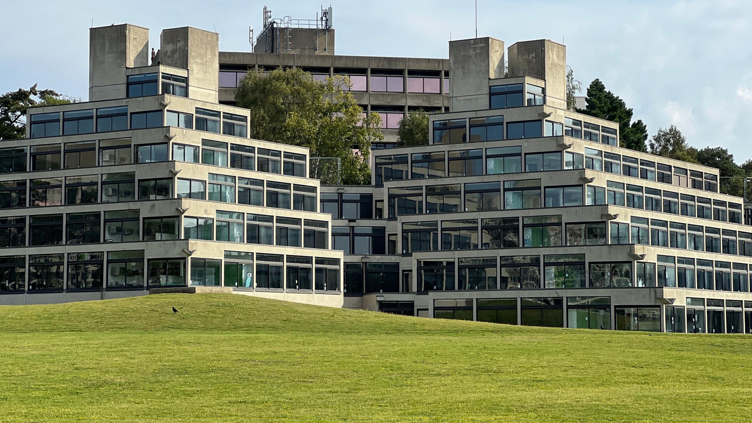 A middle-distance, modern colour photograph of the 1960s ziggurat-style halls of residence buildings at the University of East Anglia in Norwich. They are grey, brutalist 1960s buildings in the shape of stepped pyramids, with large windows running along all floors. Some more traditional grey office block-type buildings are visible behind them, and in the foreground is neatly-trimmed grass on a gentle slope up to the buildings.