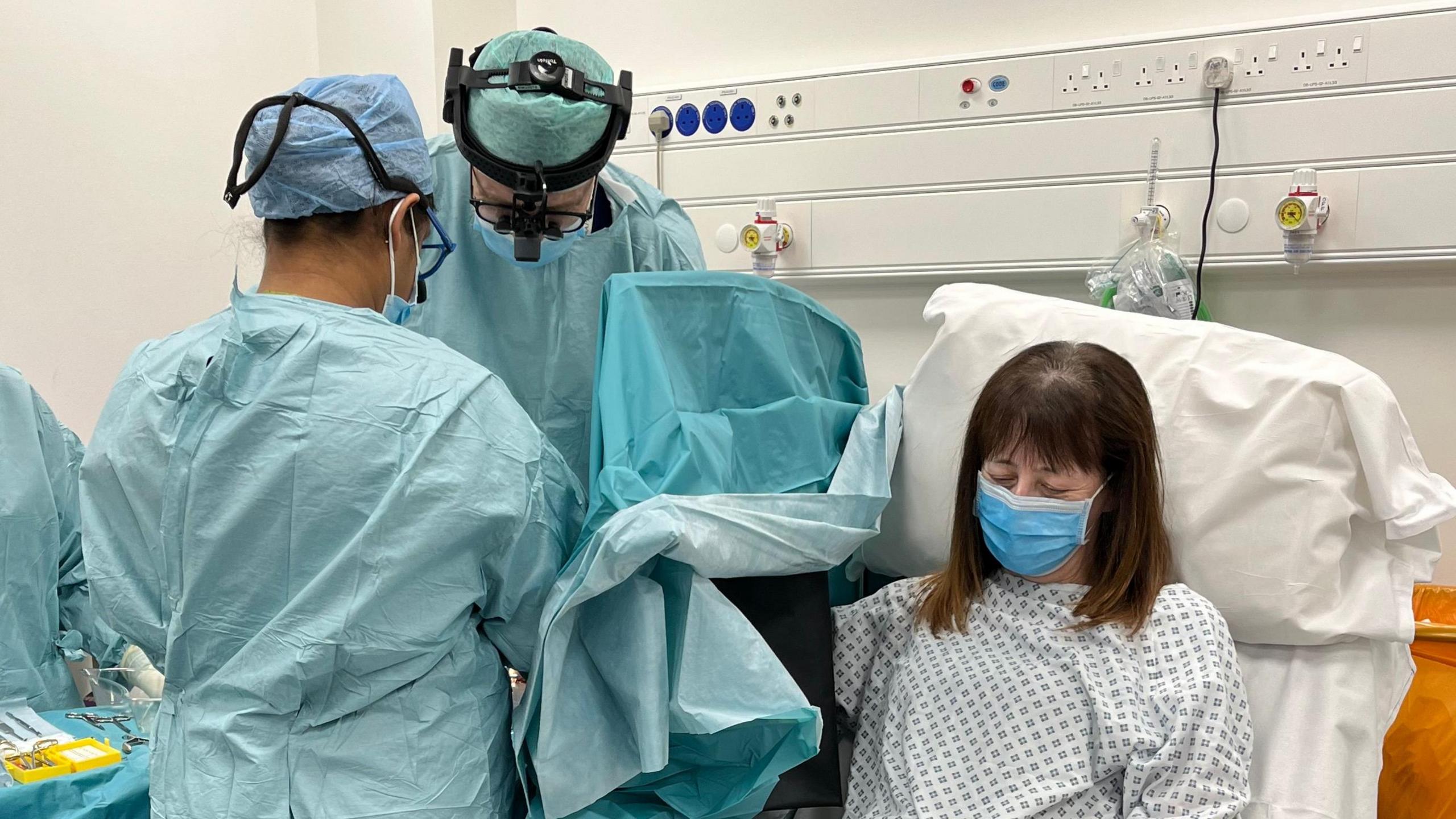 A female patient in a hospital gown with a blue face mask on sits in a chair. To the left of her, two surgeons dressed in blue surgical clothes are working on her hand, which is not visible and is behind a screen