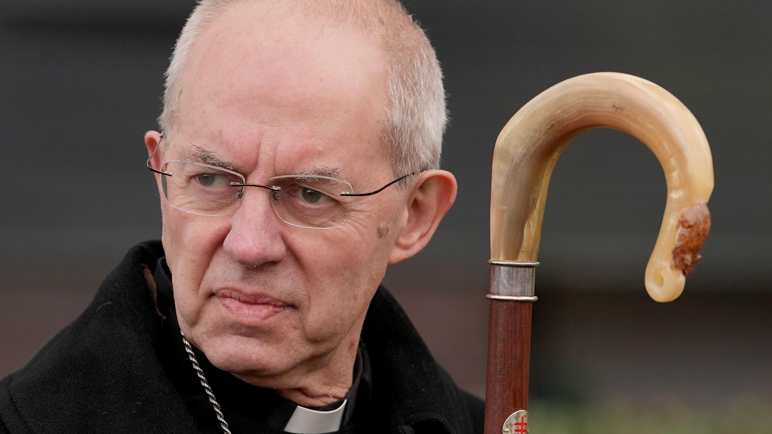 Close-up of Justin Welby, wearing glasses, clergy dog collar and holding a staff.