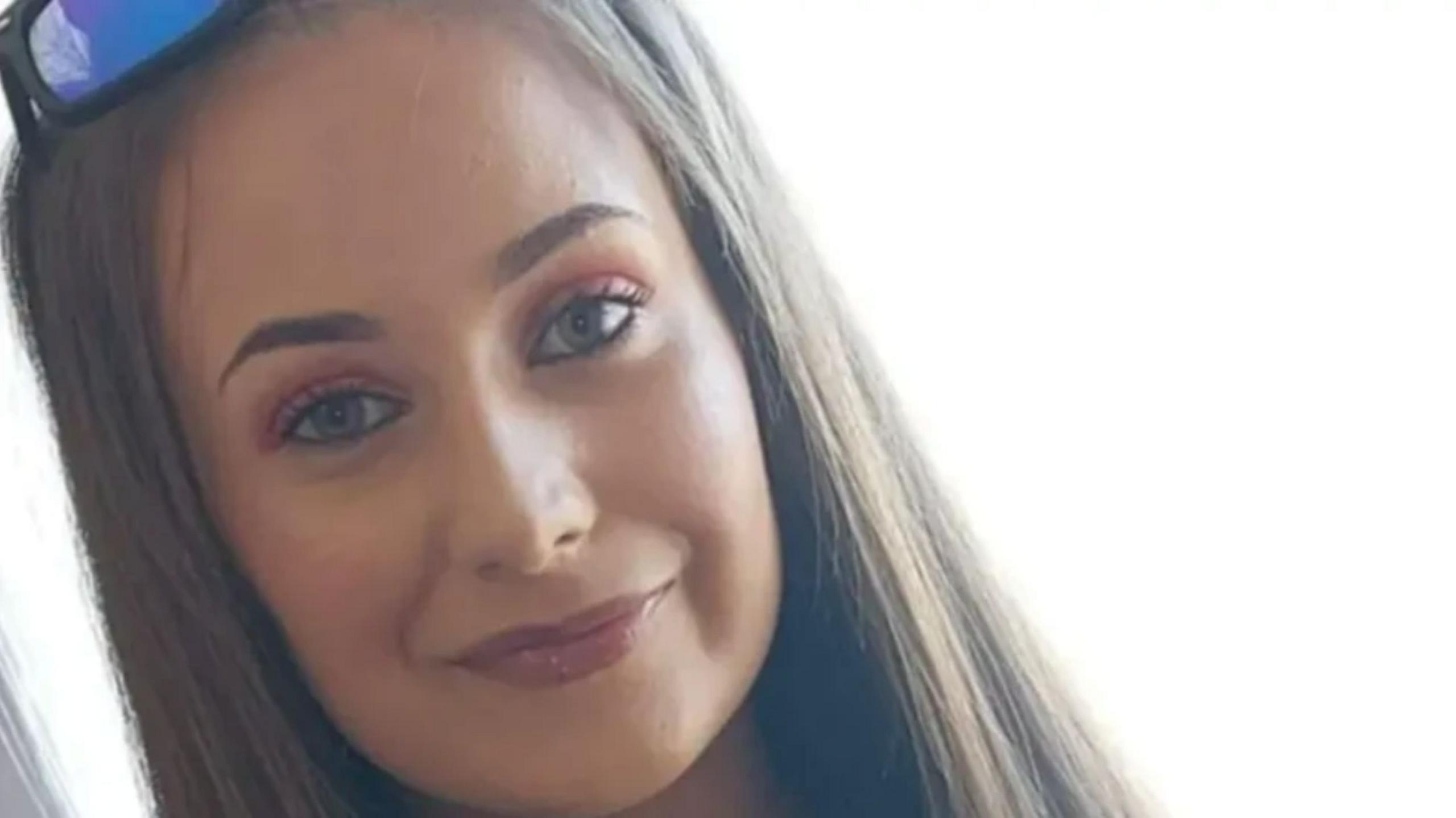 Chloe Mitchell is smiling at the camera, she has long brown hair and is wearing a pair of sunglasses on her head.
