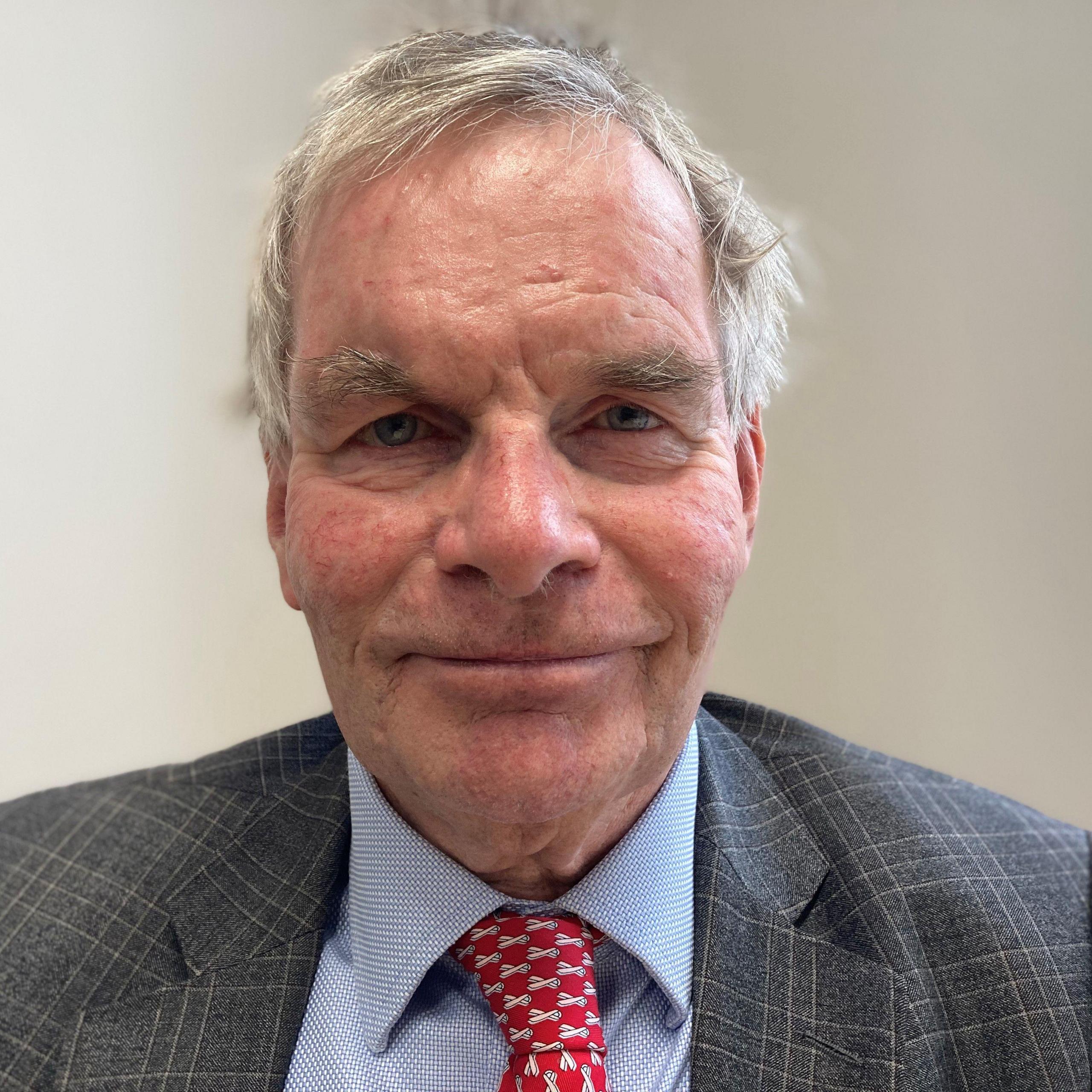 A head and shoulders photograph of Councillor Martin Hill. He has short grey hair and is wearing a grey-blue suit, blue shirt and red tie.