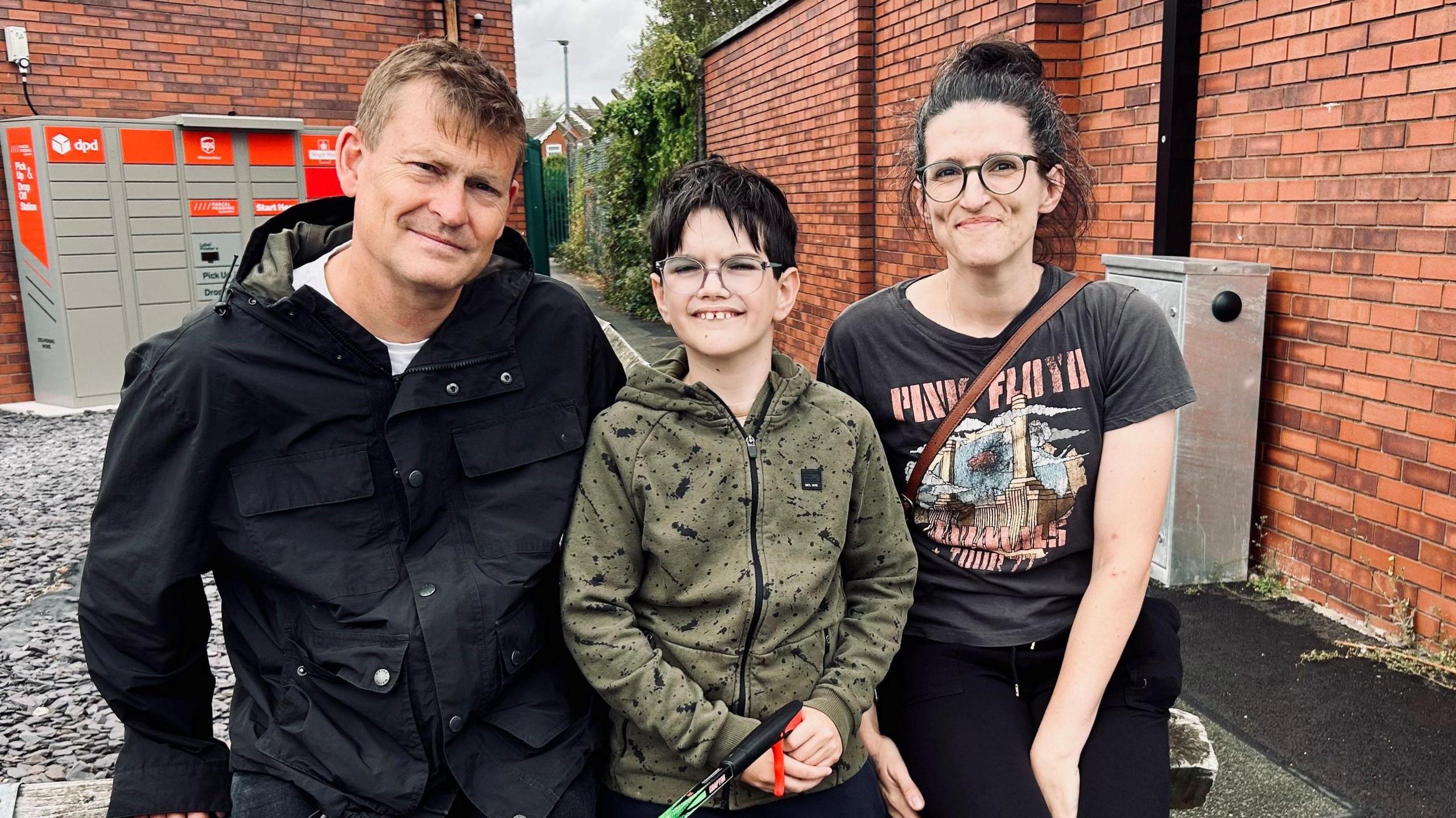 Ellesmere Port and Bromborough MP Justin Madders (left) with brown hair wearing a black coat, Jackson (centre) with black hair and glasses wearing a green hoodie and (right) Leanne with black hair in a pony tail and wearing black glasses, a black Pink Floyd T-shirt and black trousers. They are seated in front of an alleyway and all are smiling.