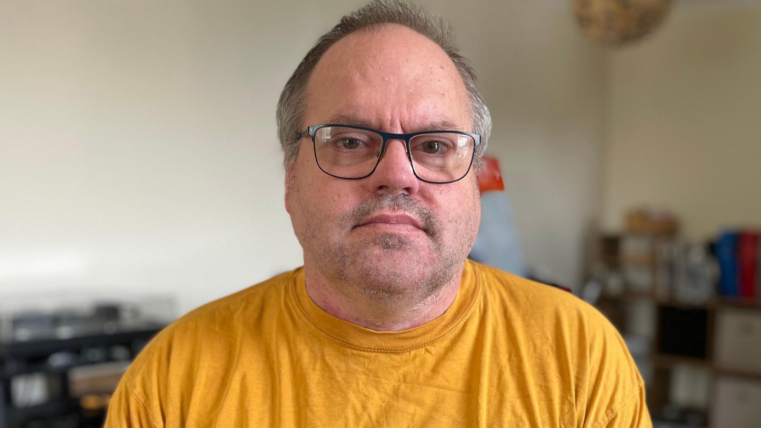A man in his fifties is looking into the camera with a neutral expression. He is sitting in his wheelchair in the lounge of his home. He is wearing a yellow t-shirt and has glasses on. 