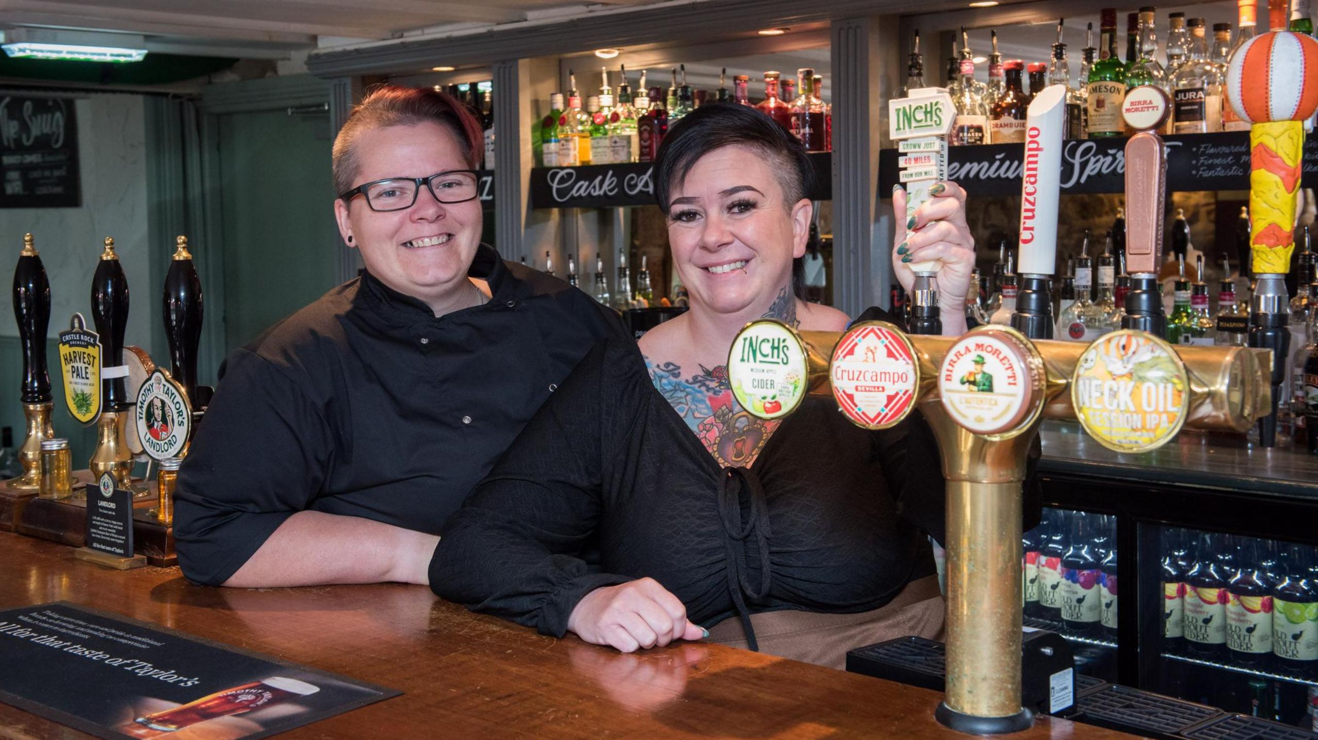 Two women stand behind a well stocked bar. They are both wearing black and smiling and one has a very colourful tattoo visible on her chest and neck. 