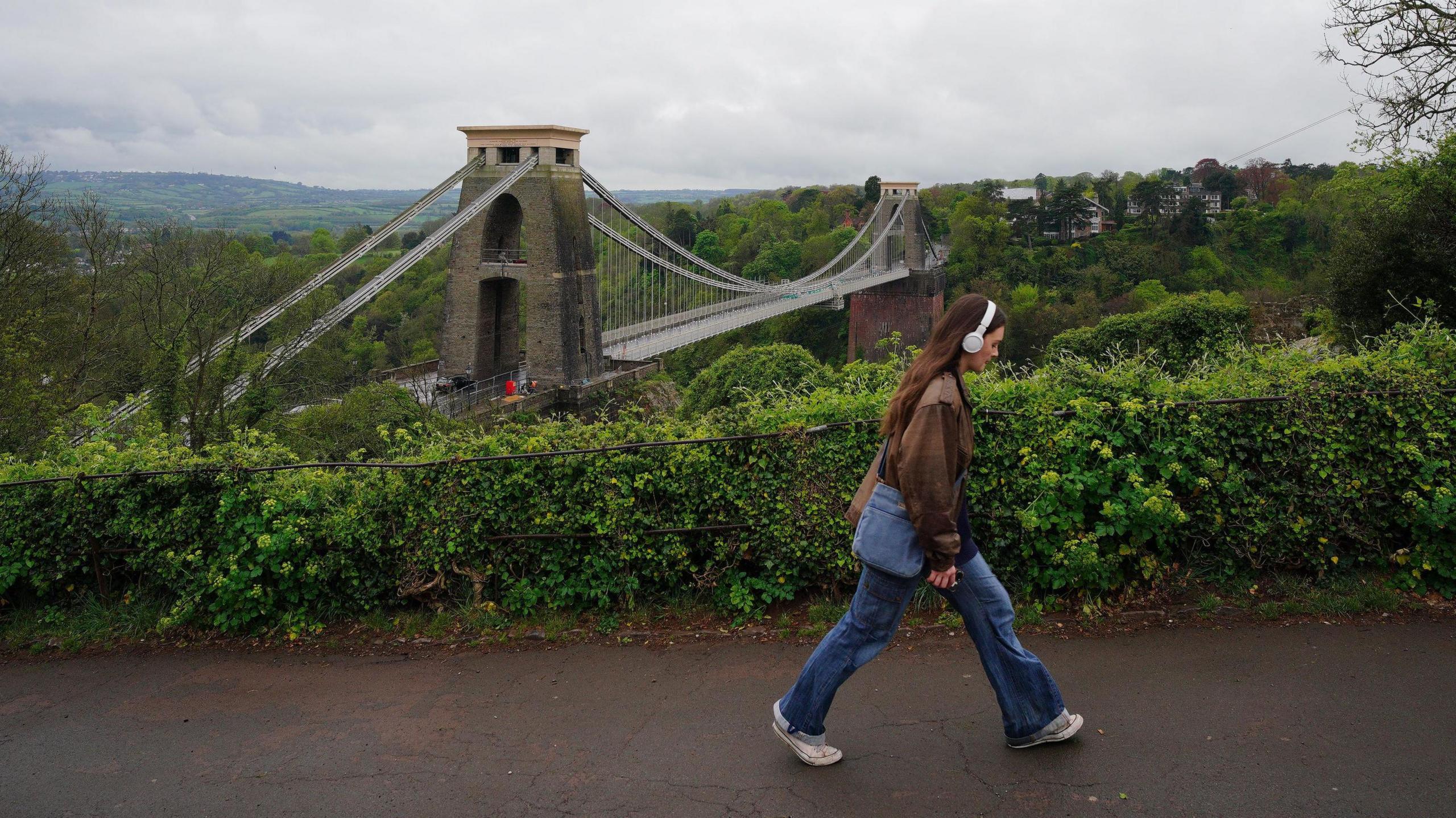 A young woman with headphones on walks near the Observatory in Bristol. In the background the Clifton Suspension Bridge is visible. It is an overcast day