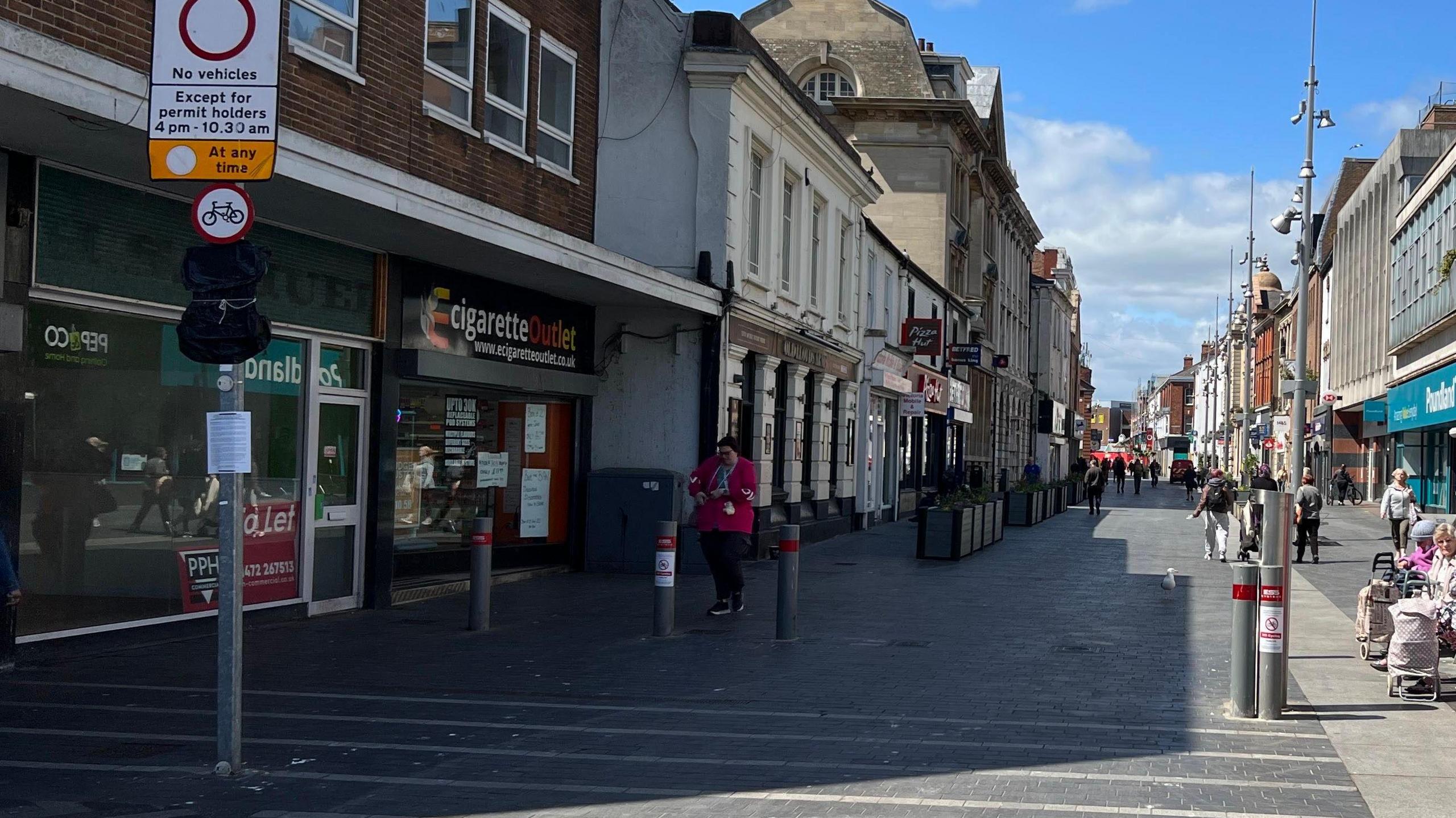 A view of Grimsby town centre shows rows of shops on either side under a blue sky. On the left, an empty store with a red "To Let" sign in the window.