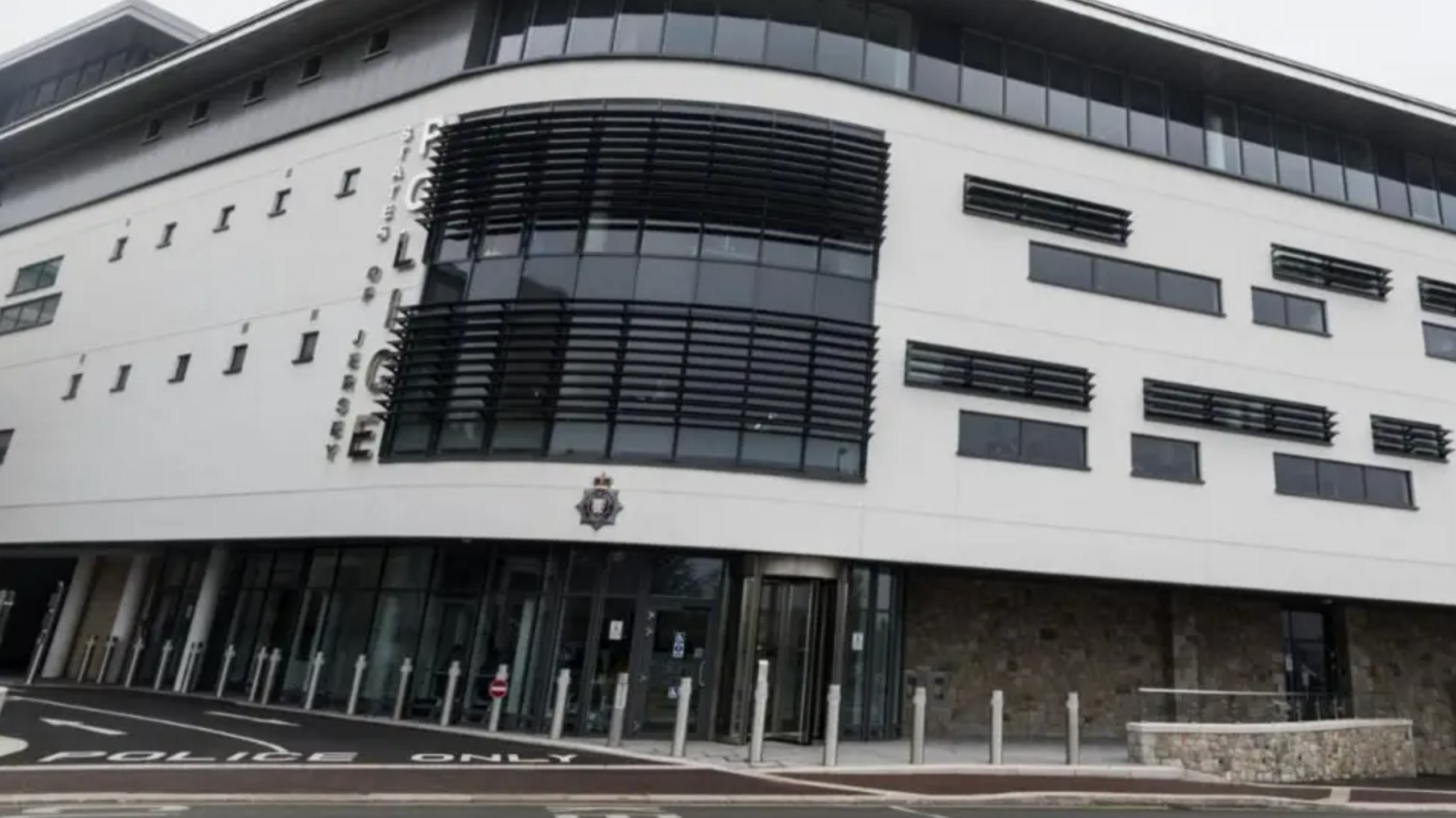 A modern multi-story police station with a curved white facade, large glass windows, and horizontal black slats. The building features a vertical 'POLICE' sign and an emblem above the entrance. The foreground includes road markings reading 'POLICE ONLY' and several bollards.