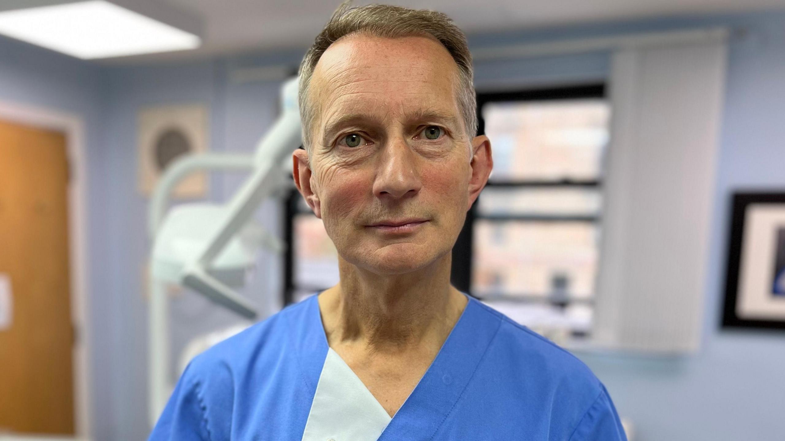 Dentist David McColl, who is wearing blue scrubs and is standing in a dental surgery, looks into the camera